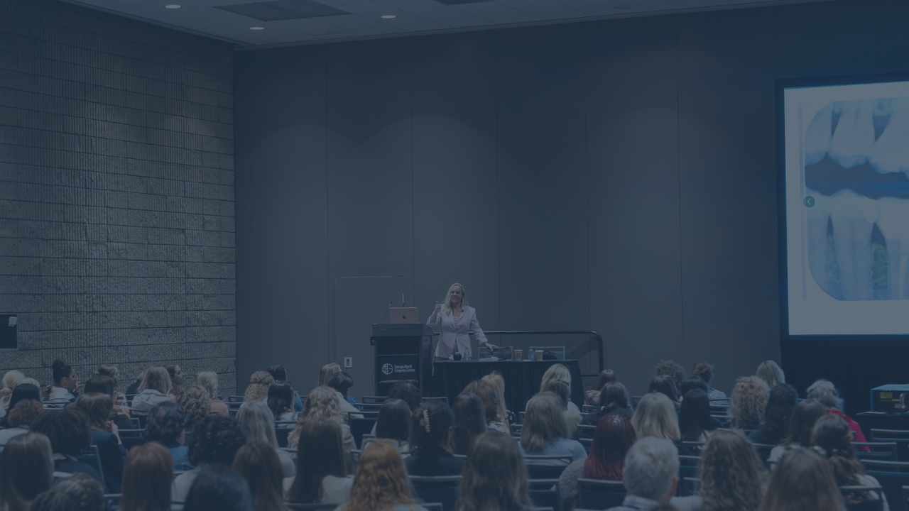 Dr. Kelly is giving a presentation to a large audience in a conference room or auditorium.