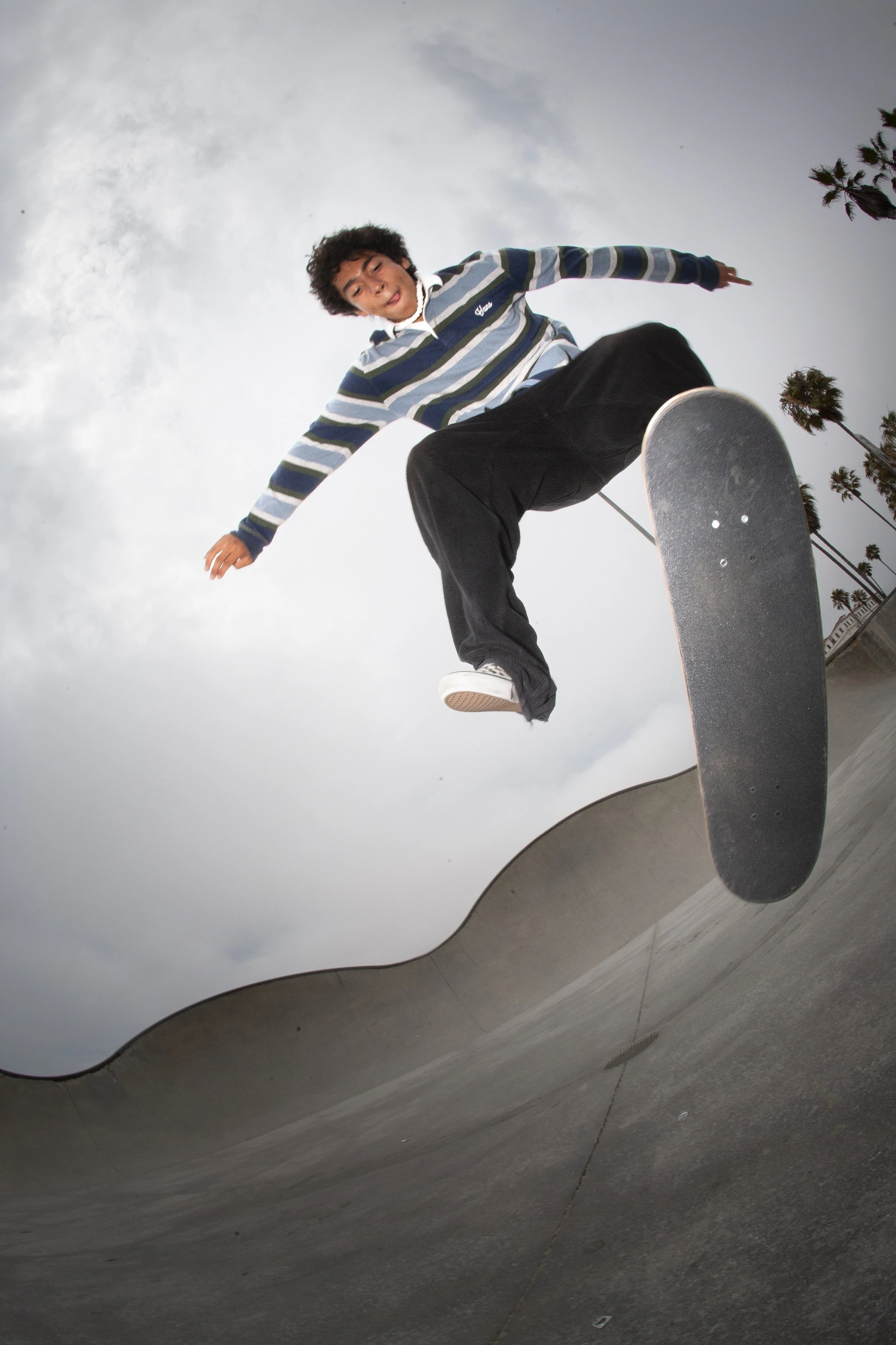 Skateboarder performing a trick at a skatepark under a cloudy sky with palm trees in the background.
