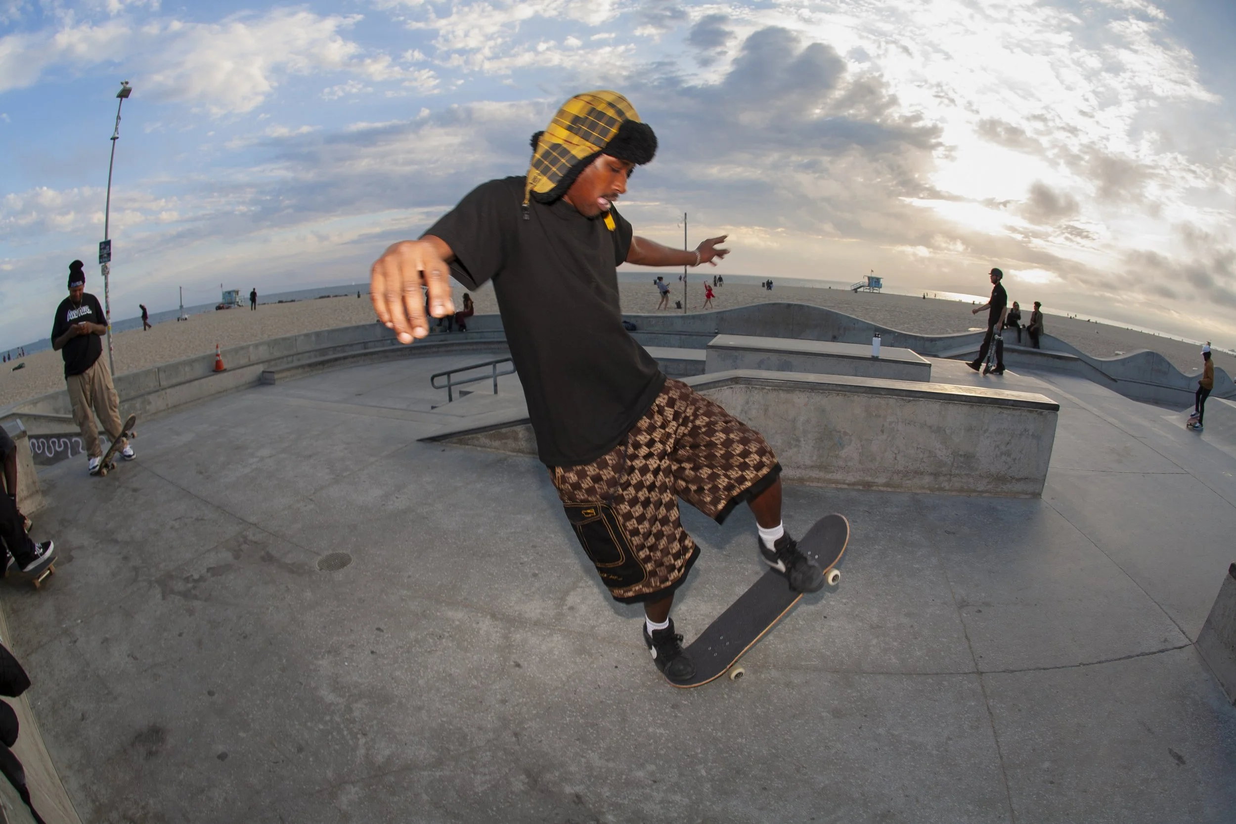 Skateboarder performing a trick at a skate park near the beach during sunset with other skaters and people in the background.