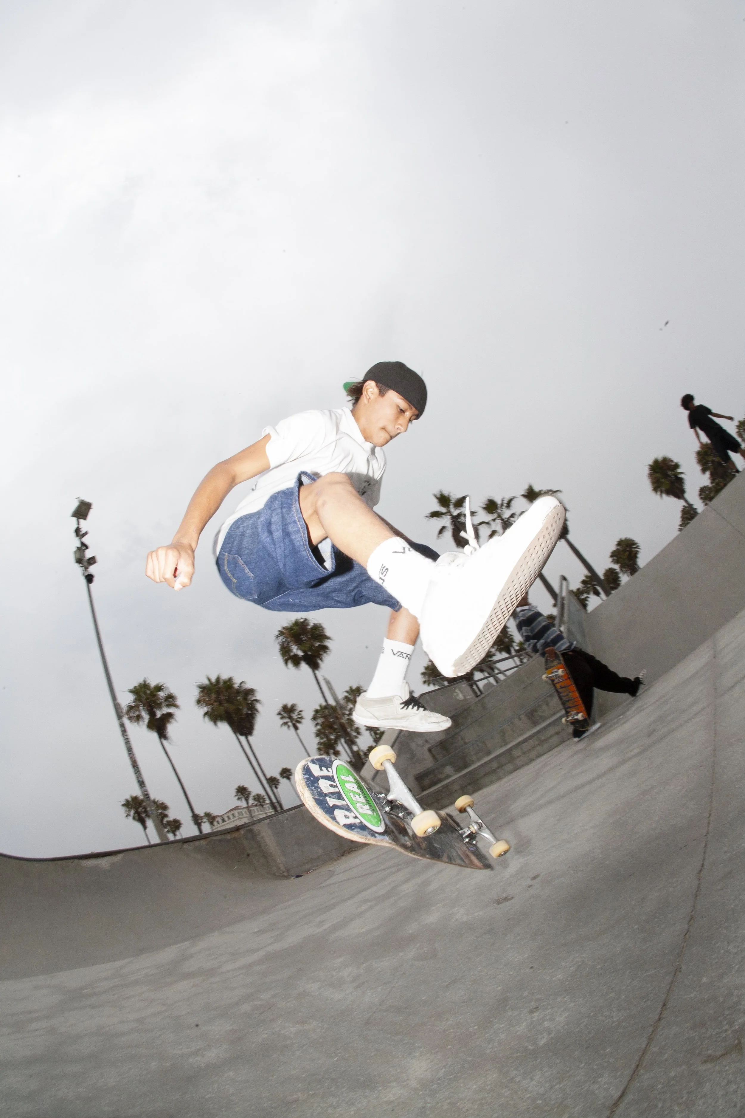 Skateboarder performing a trick at a skatepark with palm trees in the background under a cloudy sky.