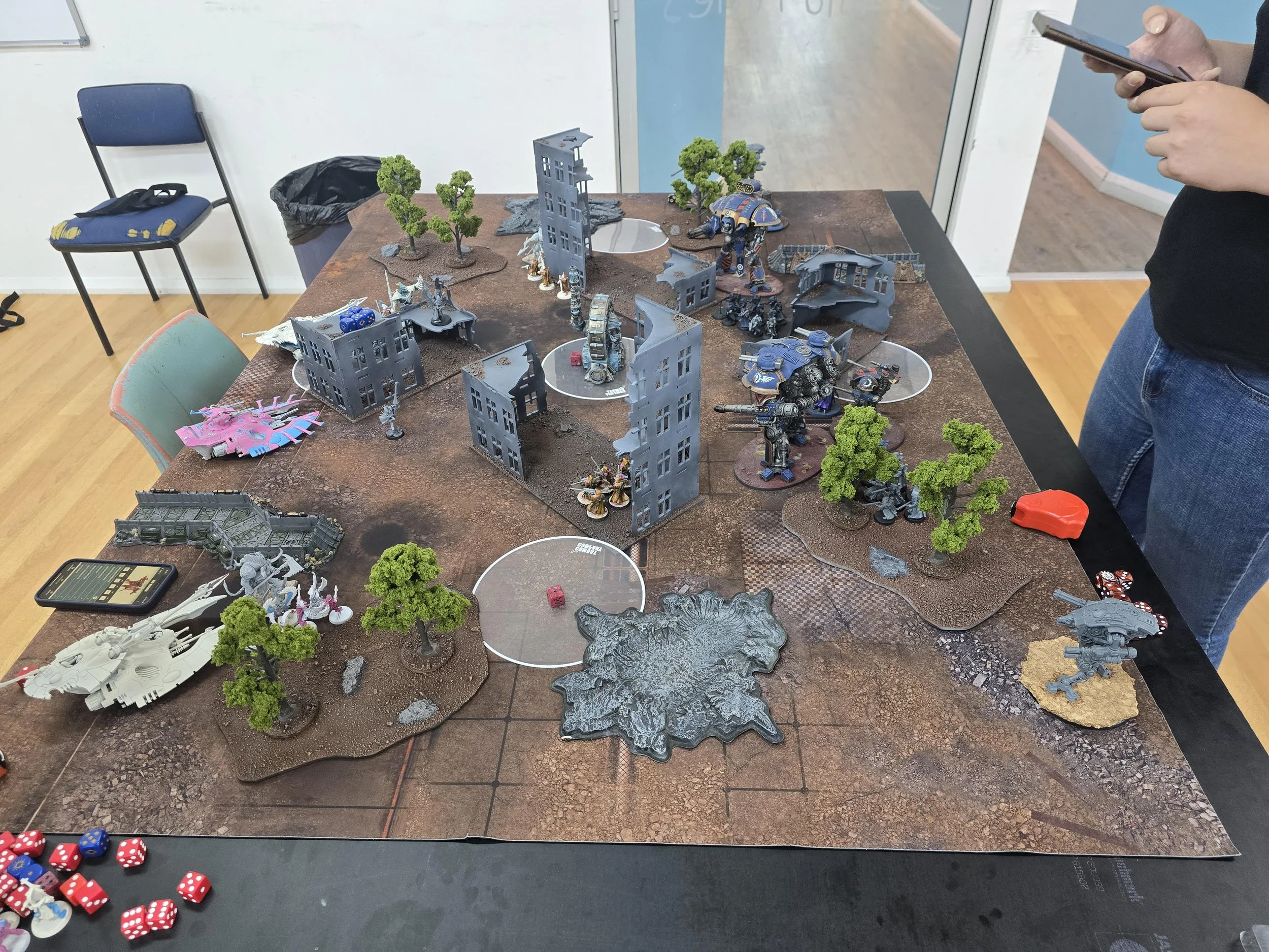 Tabletop wargame with miniature figurines, vehicles, dice, and terrain on a gaming board.
