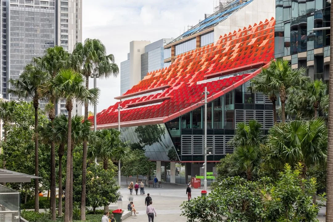 Modern building with a red and orange geometric roof surrounded by palm trees and pedestrians.