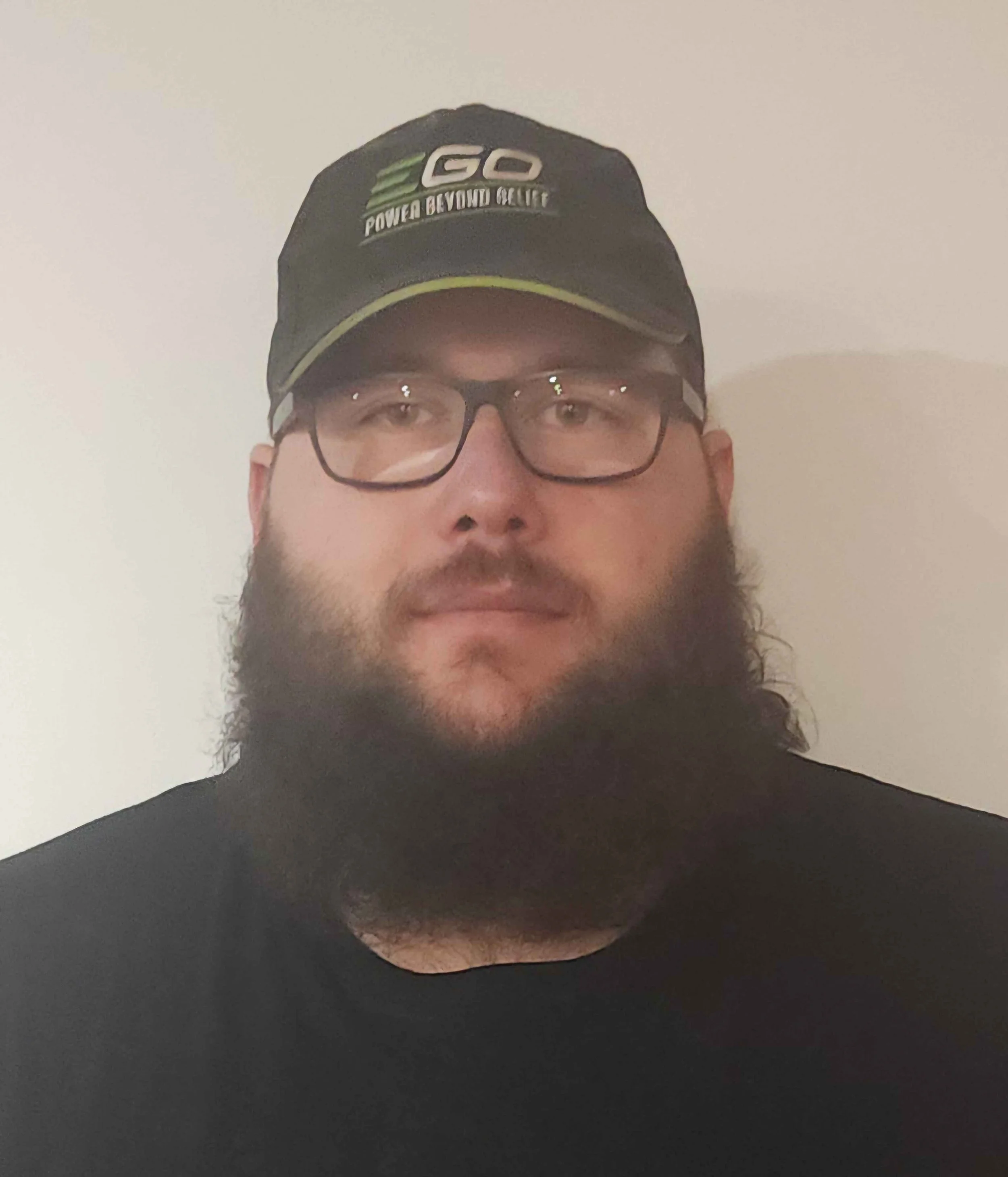 A man with glasses, a full beard, and long hair wearing a black cap and black shirt standing against a plain background.