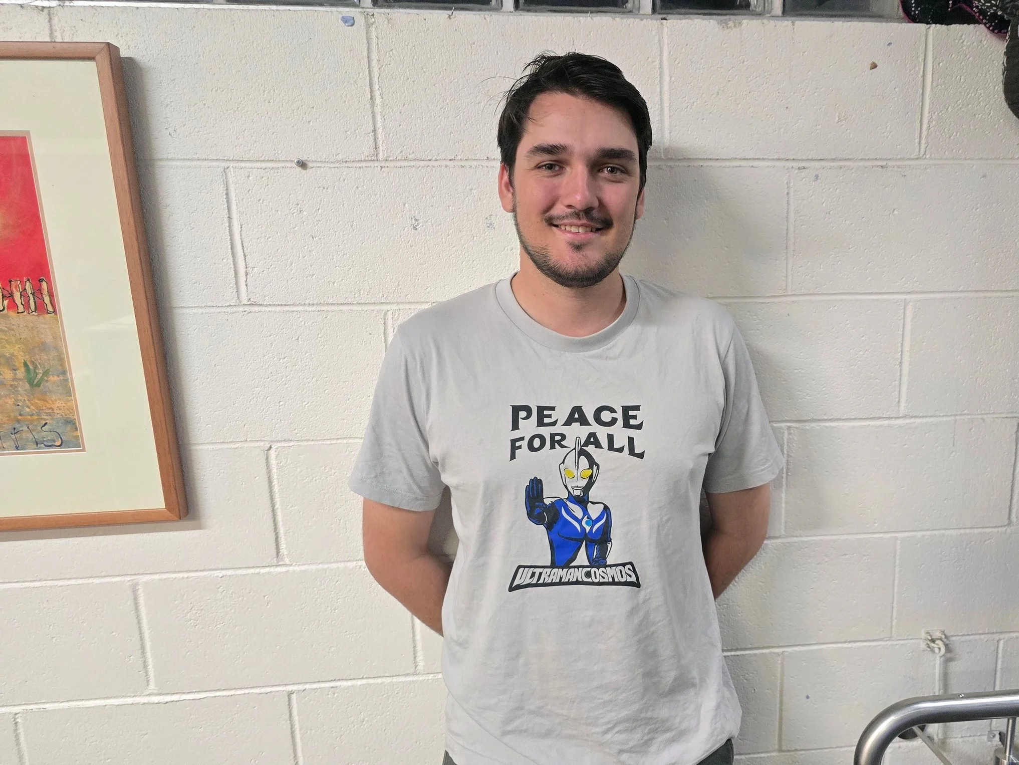A young man with dark hair and a beard smiling, standing against a white brick wall. He is wearing a light gray T-shirt with a graphic of a robot and the words "Peace for All" and "Ultraman Cosmos".
