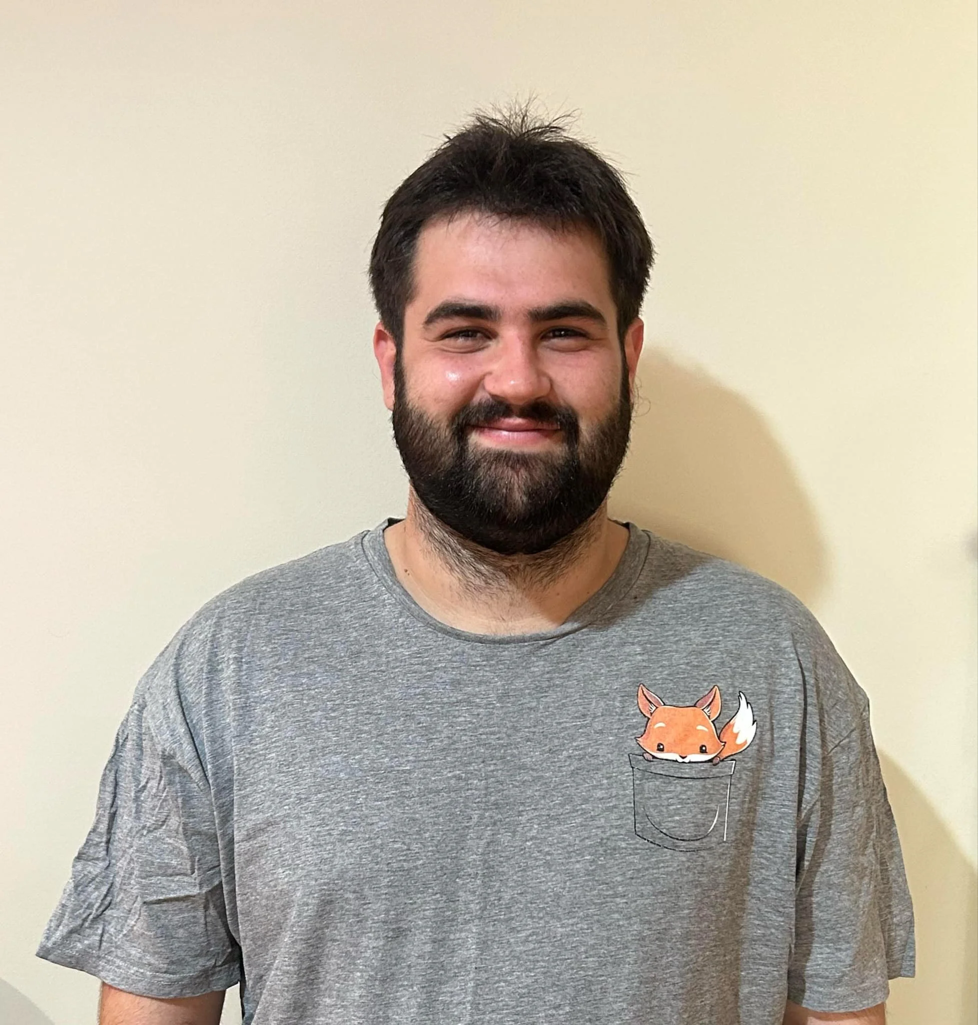 A man with dark hair and beard smiling, wearing a gray t-shirt with a fox graphic on a small pocket.