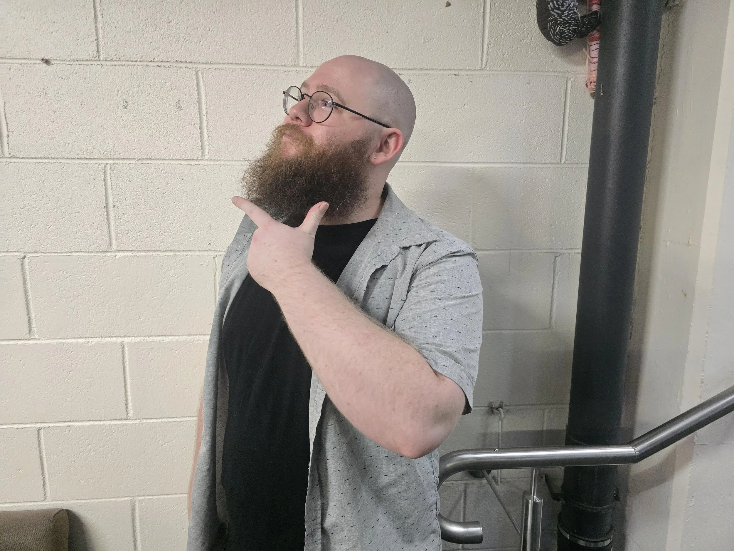 Bald man with glasses and a beard, wearing a black shirt and a light gray shirt, standing in front of a beige cinder block wall, posing with his right hand under his chin and looking upward.