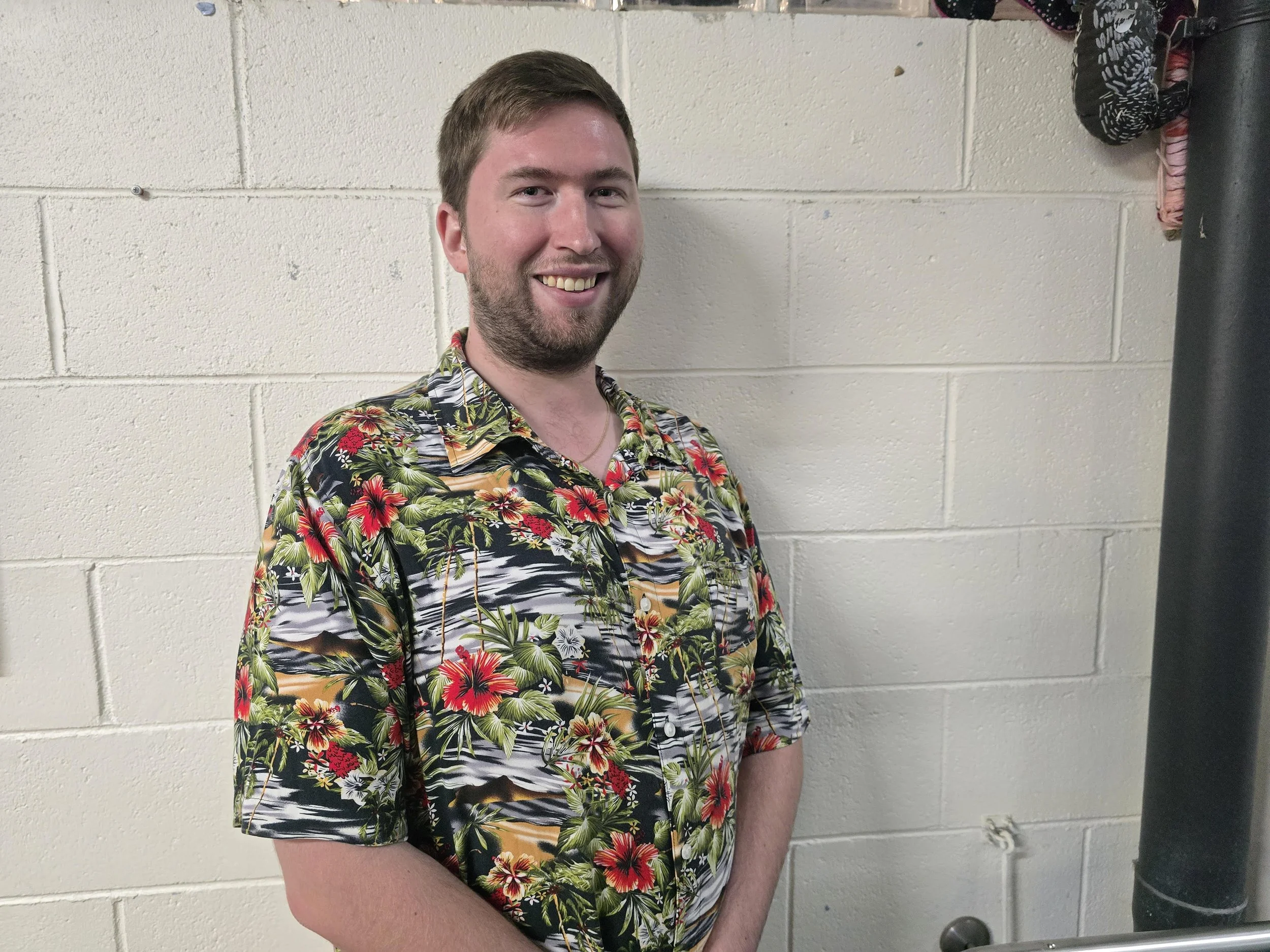 A man with light brown hair and a beard, smiling, wearing a colorful Hawaiian shirt, standing against a white brick wall.