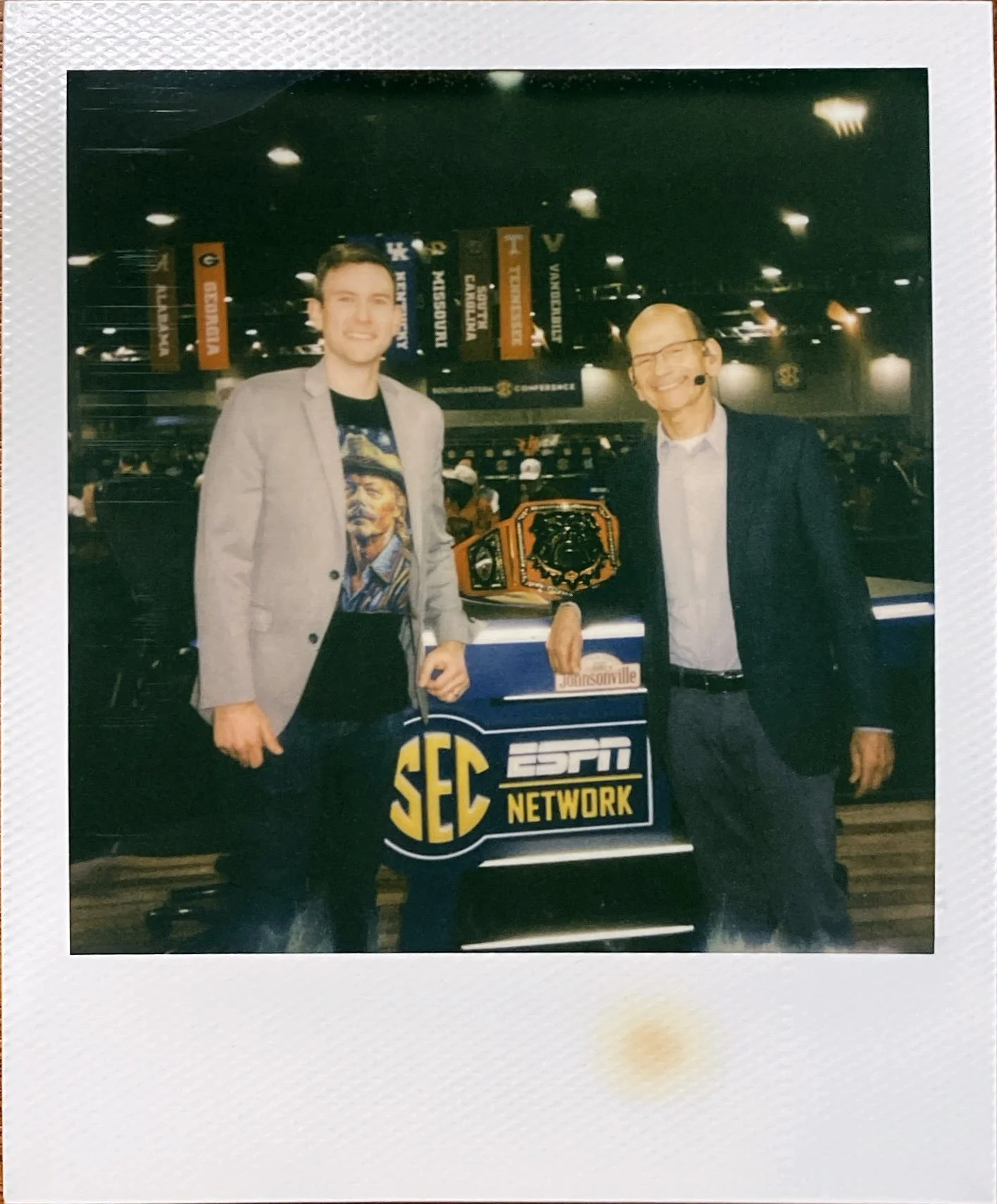 Two men standing in an indoor arena with banners hanging from the ceiling. One man is wearing a gray blazer and a graphic t-shirt, and the other is wearing a dark blazer with a microphone headset. Between them is a display with a championship belt and a sign with the SEC and ESPN Network logos, indicating a sports or conference event.