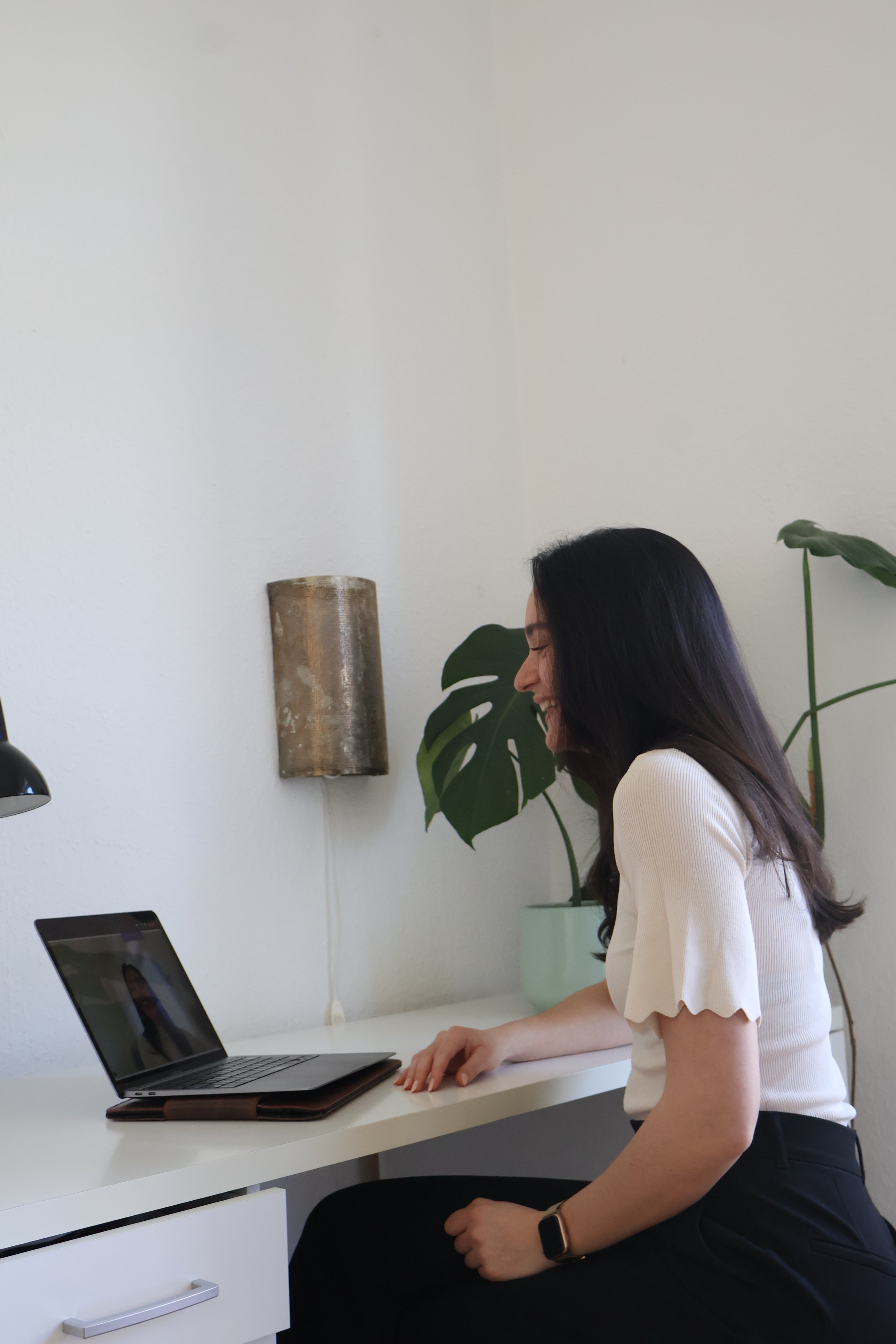 Eine Frau sitzt an einem weißen Schreibtisch, spricht in ein Laptop, trägt eine weiße Bluse und eine smartwatch, im Hintergrund sind Pflanzen und eine Wandlampe.
