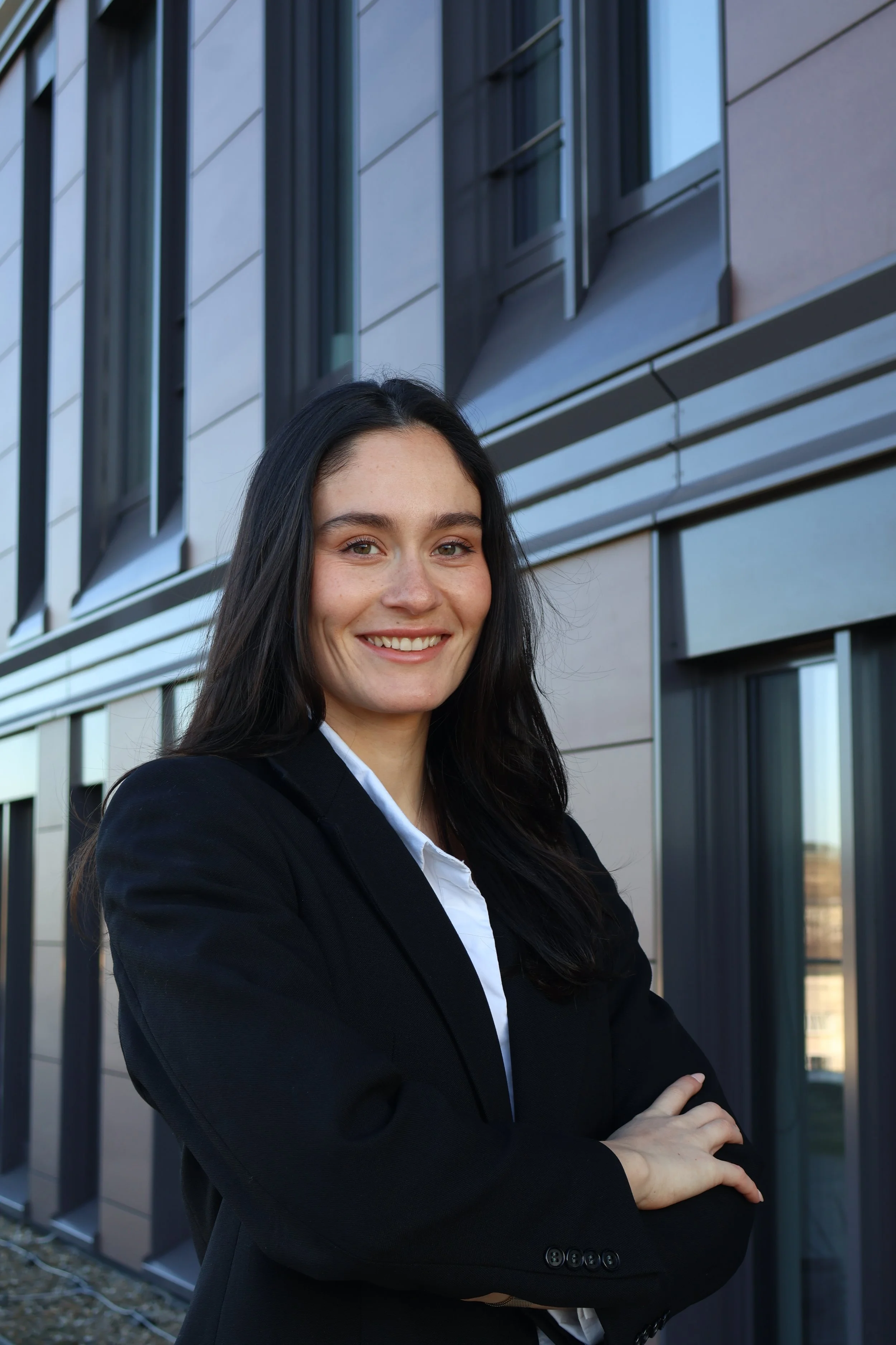 Junge Frau in Businesskleidung vor modernem Bürogebäude mit Fassade aus Glas und Metall, lächelnd bei Tag.