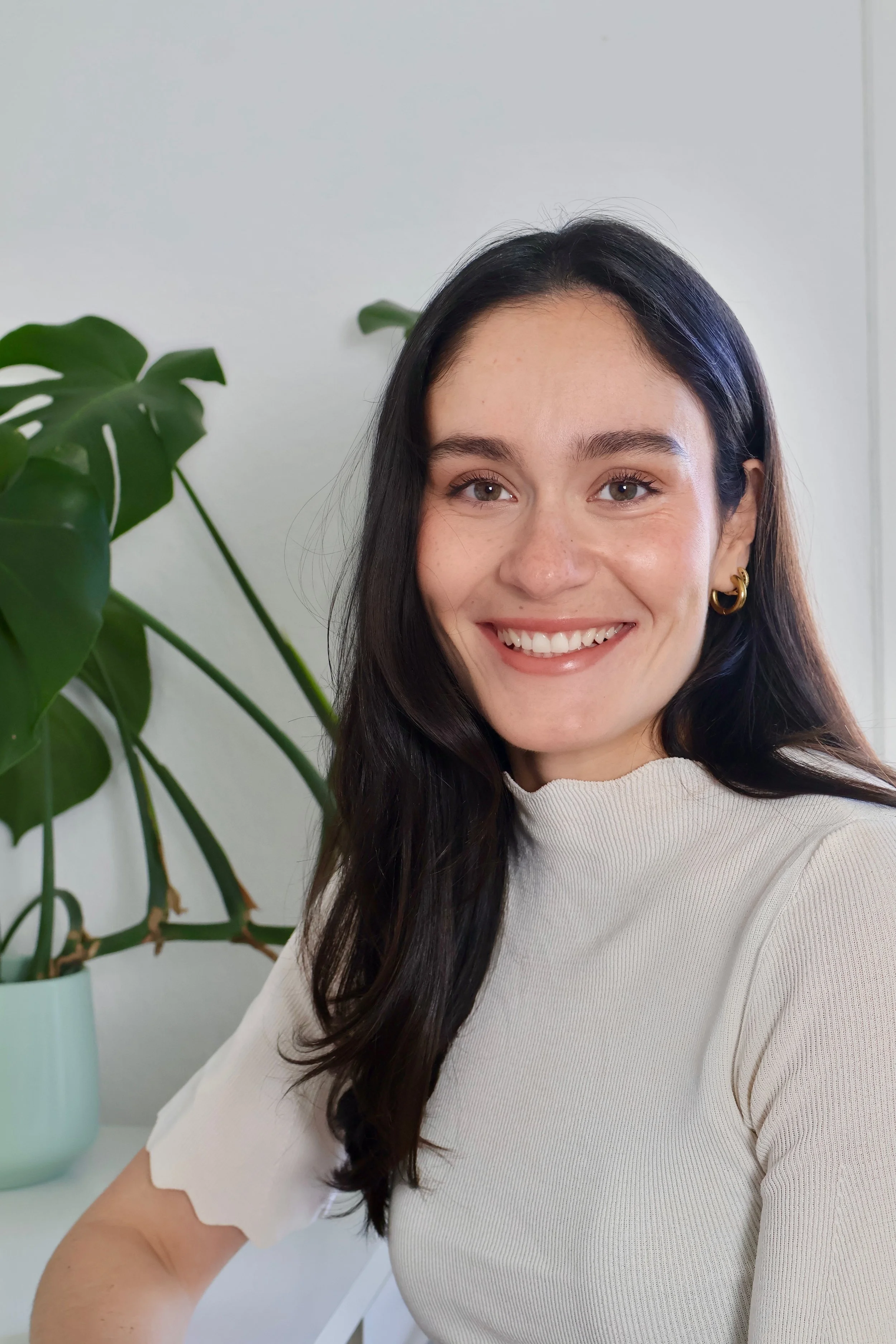 Eine junge Frau mit dunklen Haaren, die einen weißen Pullover trägt, lächelt vor einer Pflanze mit großen grünen Blättern auf einem weißen Tisch.