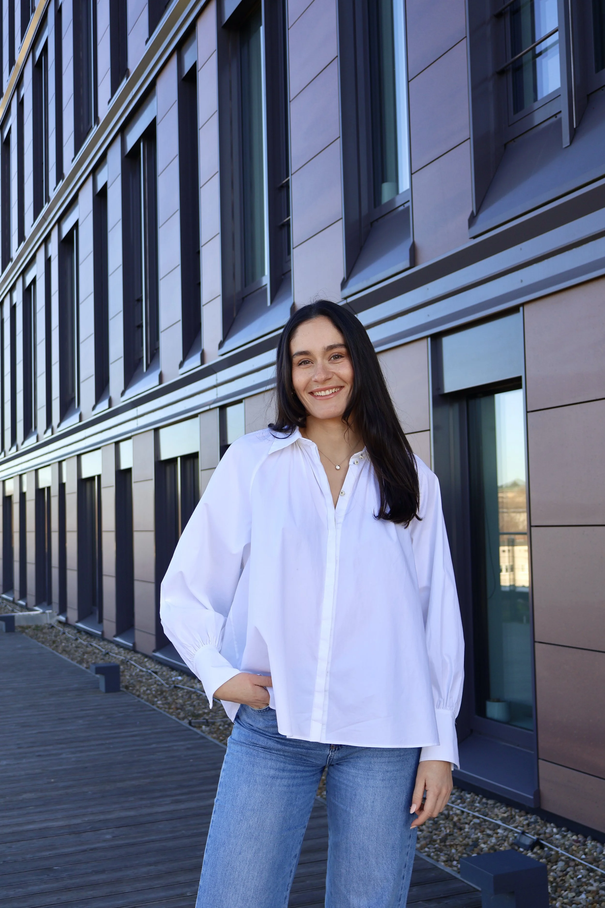 Junge Frau mit dunklen Haaren, weißer Bluse und blauen Jeans steht vor einem modernen Gebäude mit großen Fenstern.
