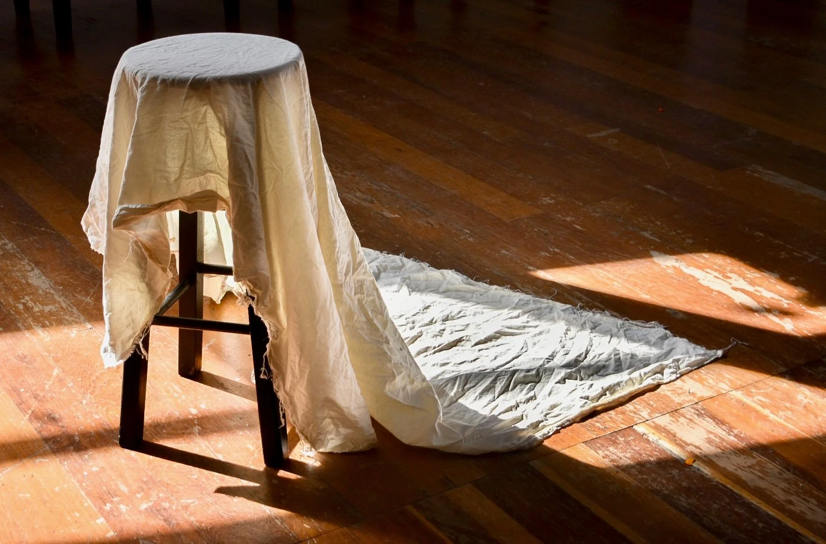 A wooden chair with a draped white cloth on top, in sunlight, with crumpled fabric on the floor next to it.