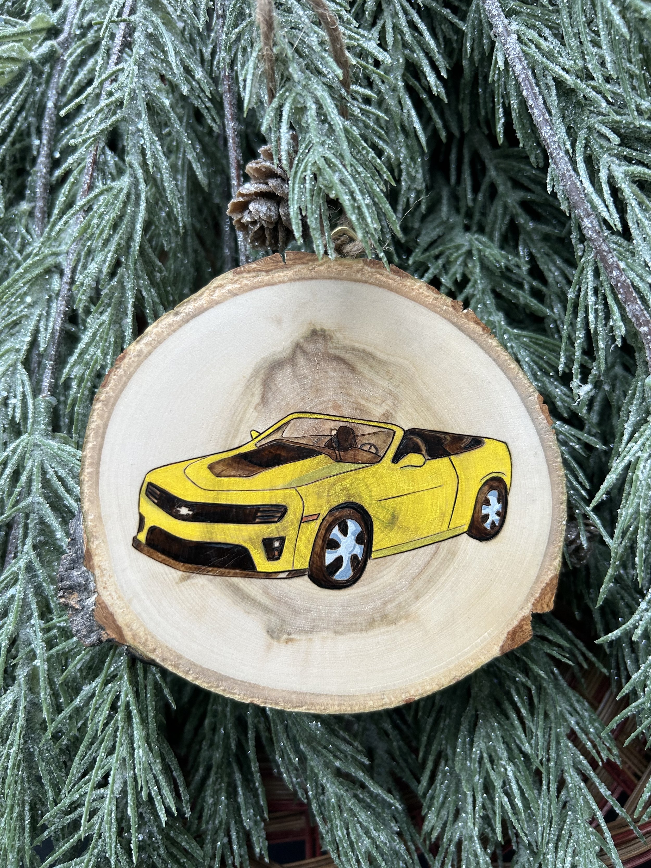 A yellow Camaro is woodburned and painted onto a small slice of Aspen wood to make a Christmas ornament. The ornament is shown hanging amid evergreen needles as if on a Christmas tree.