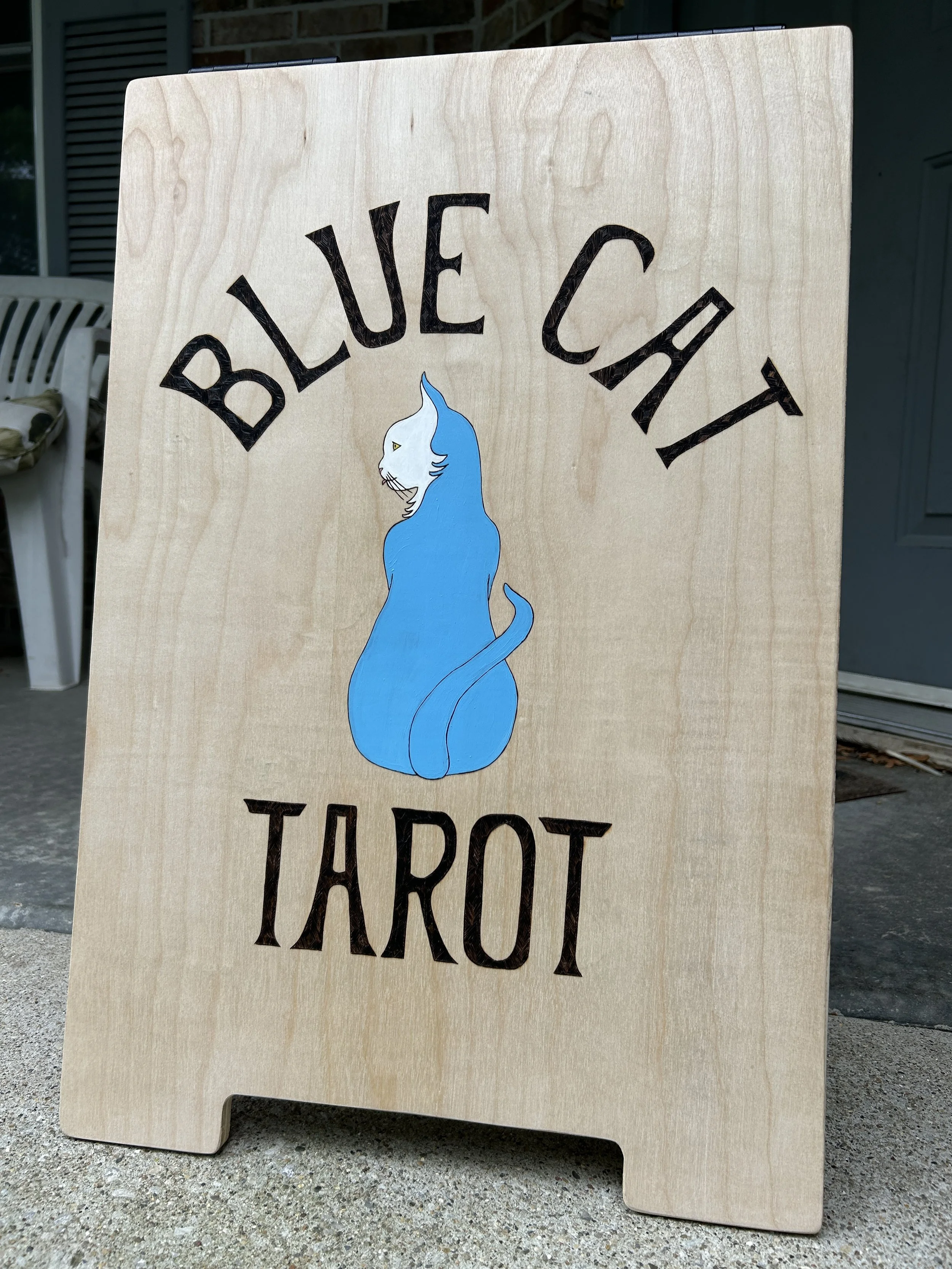 A wooden sandwich board sign reading "Blue Cat Tarot" sits on a front porch. A stylized blue cat is painted in the middle, back to the viewer and head turned to the left.