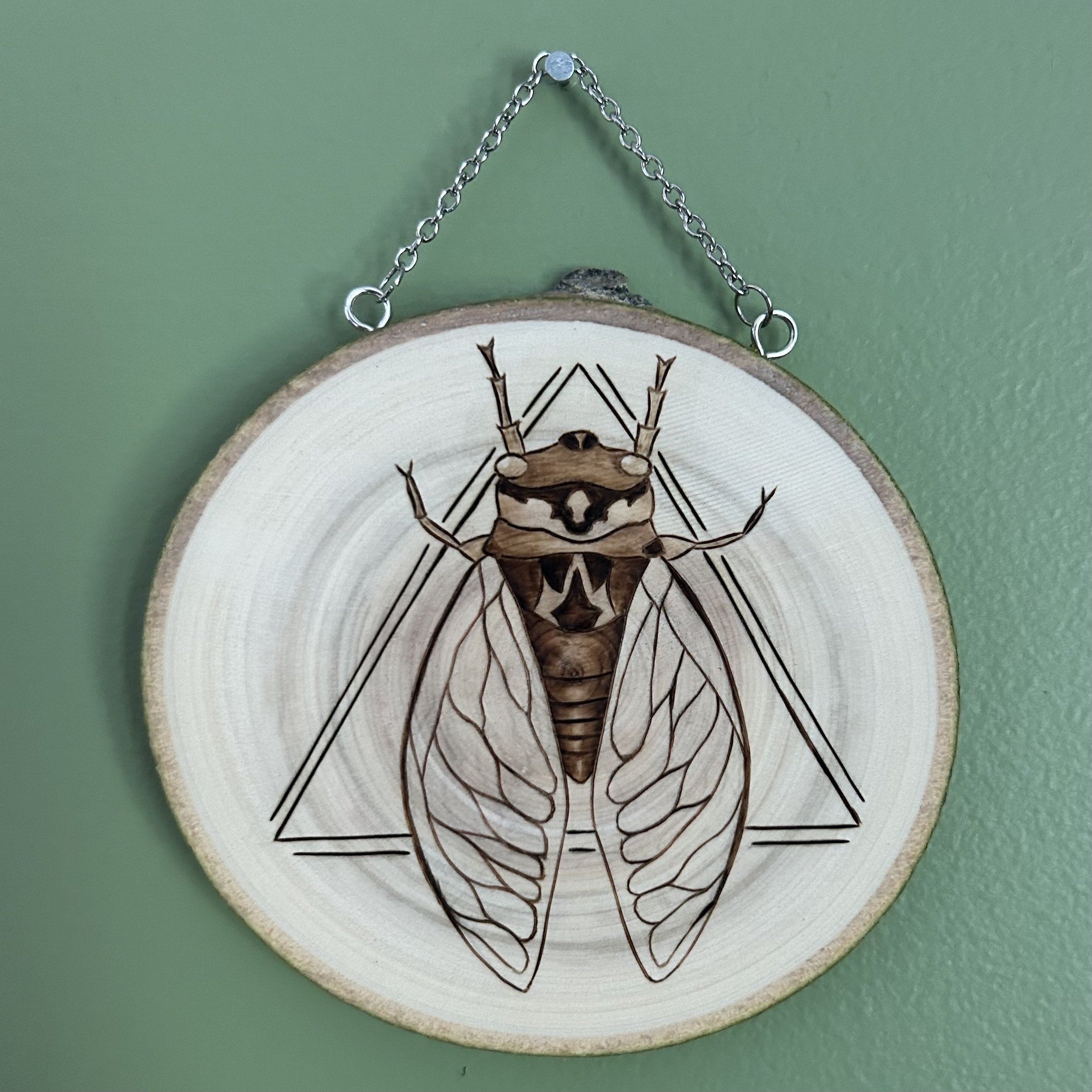 An image of a cicada superimposed over two triangles is shown woodburned onto a slice of Aspen wood hanging from a chain.