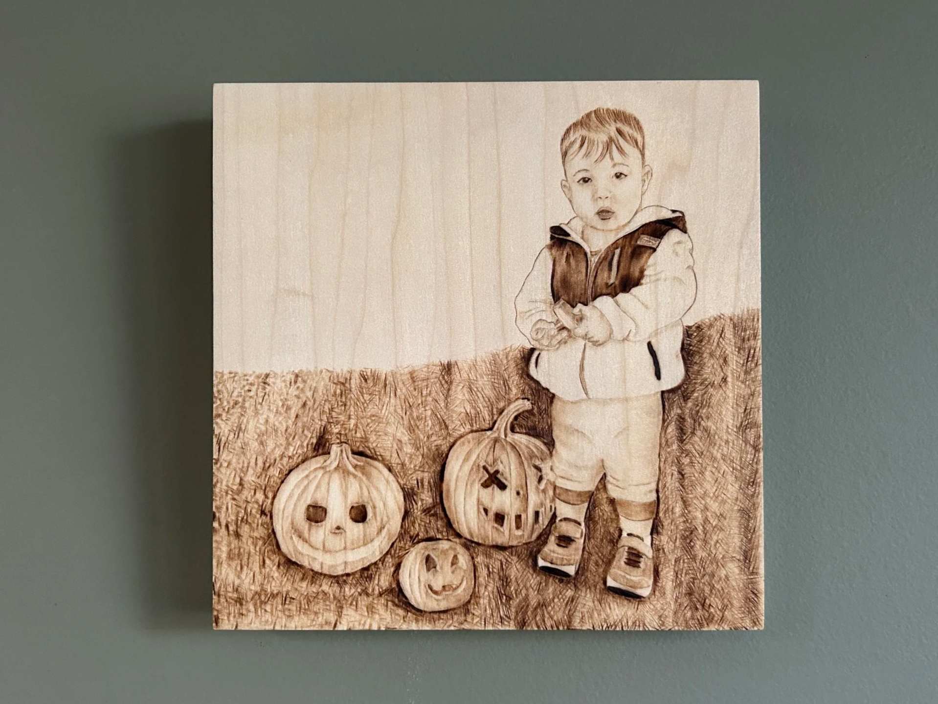 A woodburned portrait of a young boy standing next to three jack-o-lanterns
