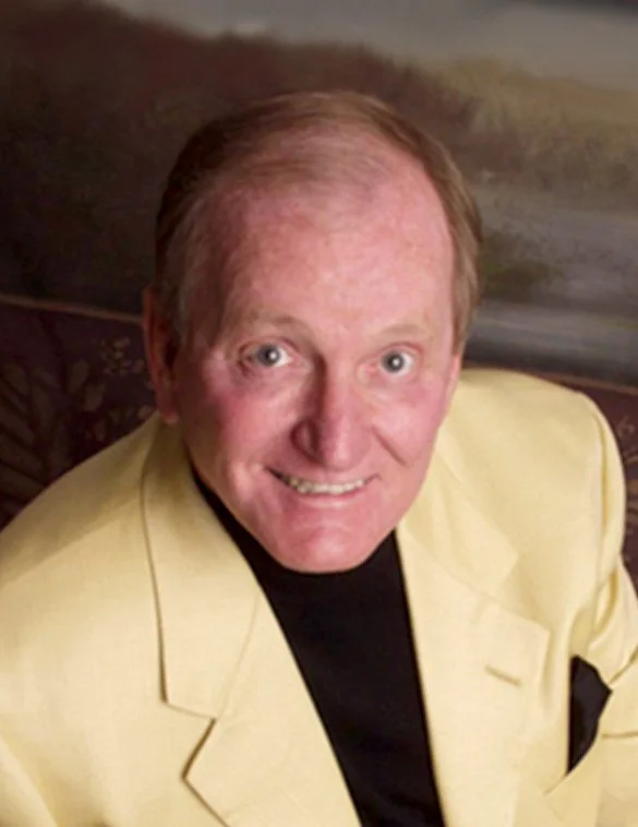 A man with short, light-colored hair wearing a dark suit, white shirt, and patterned tie, smiling at the camera against a neutral background.