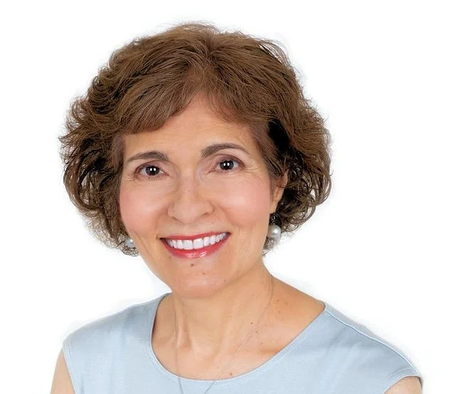 Portrait of a smiling woman with short brown hair, wearing a light blue sleeveless top and a pearl earring, against a white background.