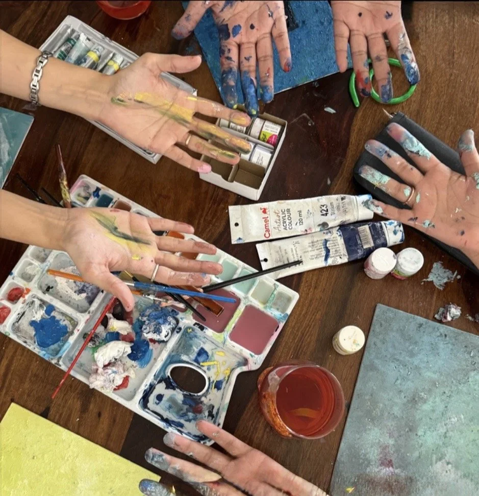 Art therapy class, there are hands in a circle facing palms up, covered in paint, above a workspace full of paint palettes and paint tubes