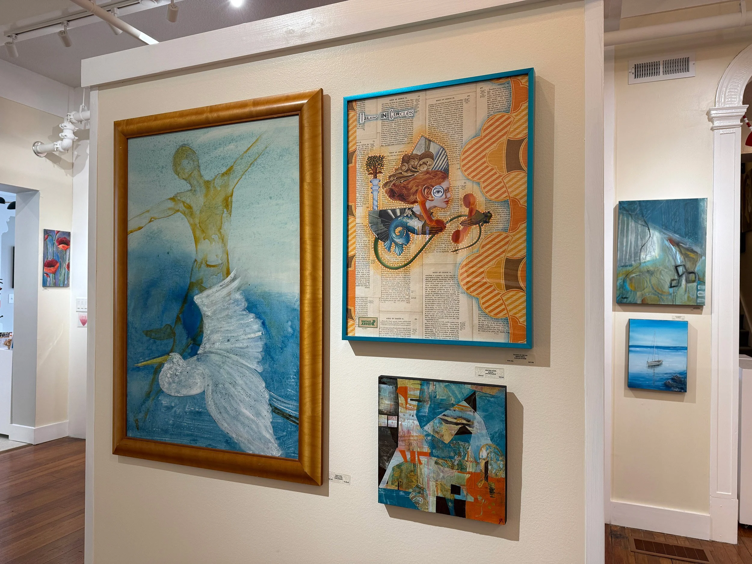 Gallery wall with five paintings. The prominent one depicts a ballet dancer with a bird in a maritime setting. The second features a stylized face with orange hair and airplanes on a background of newspaper clippings. Smaller paintings include an abstract piece with orange and blue tones, a sailboat on water, and an abstract scene with trees or branches.