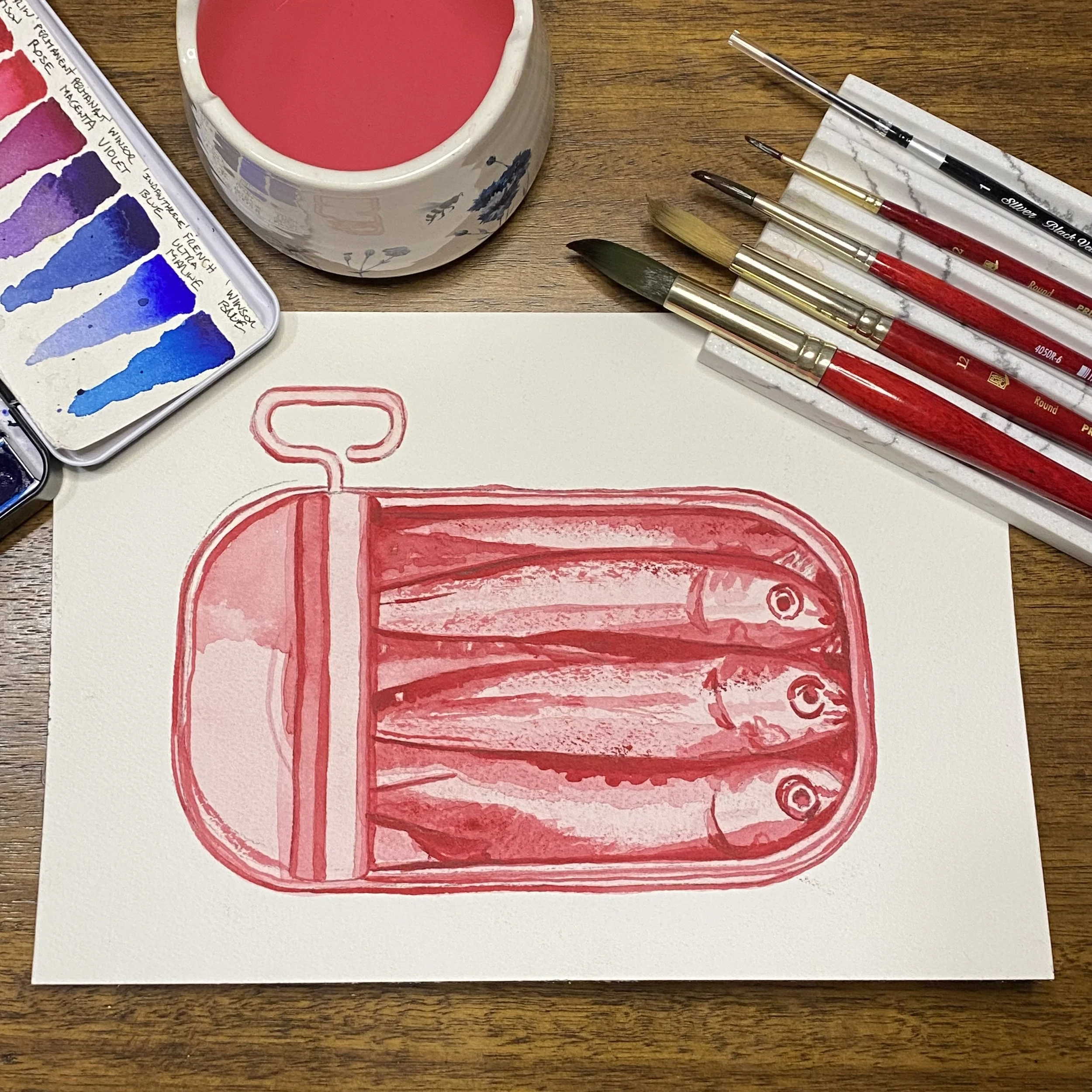 Paints and paint brush styled with a painting, in red watercolor, of a charming tin of sardines.