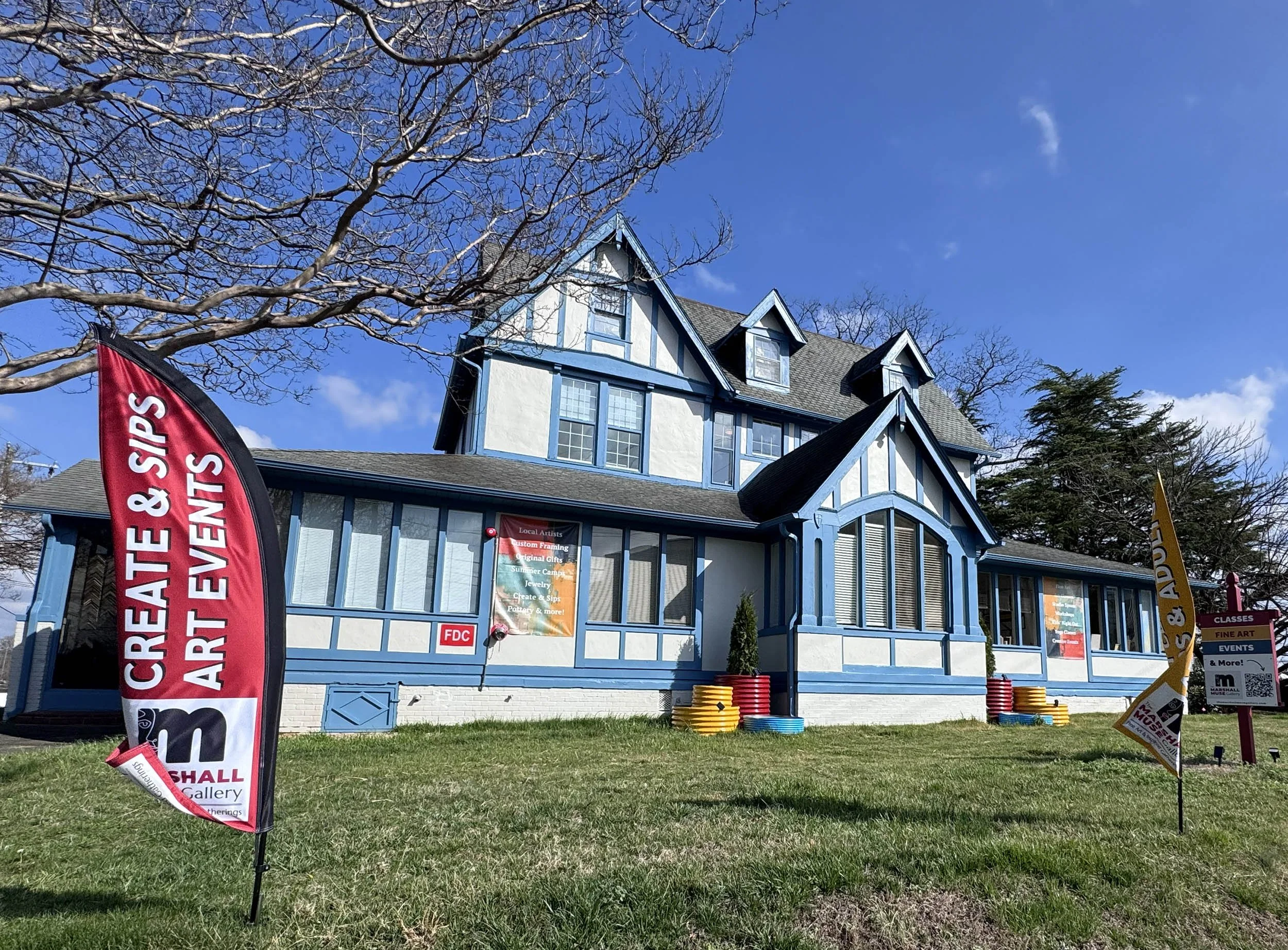 The Marshall Muse Gallery, an 8700 square foot Tudor-style home in downtown Greensboro. Cream with blue trim, brightly colored signs and planters in red, blue and bright yellow. The bright blue sky and lush green grass offset the vivid shot.