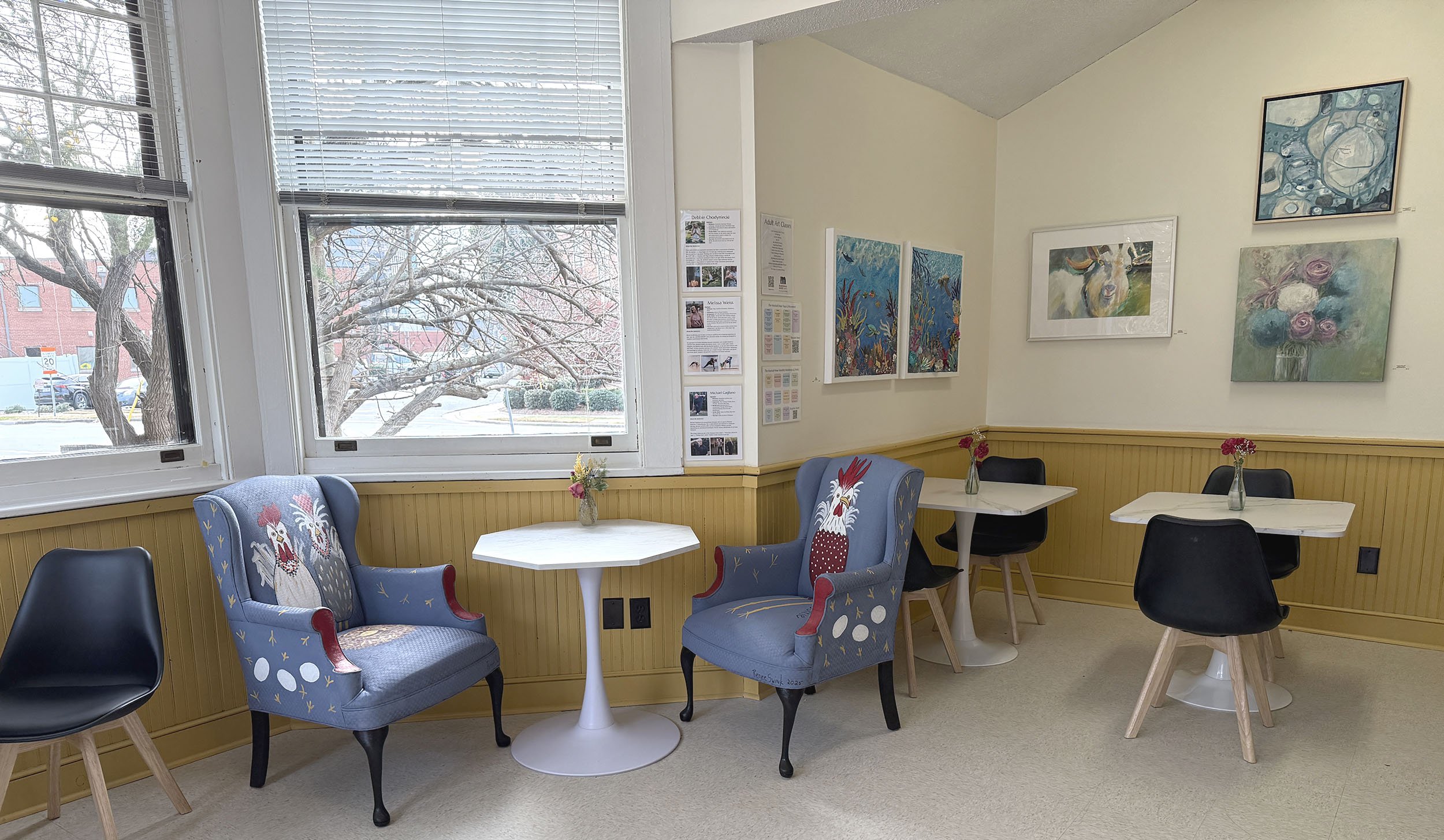 A cozy indoor cafe or lounge with several black and wooden chairs around white tables. Two blue armchairs with rooster embroidery on the backrests face each other near a window, which shows snow-covered trees outside. The walls are decorated with colorful artwork and informational posters. Small vases with pink and yellow flowers sit on the tables, creating a welcoming atmosphere.