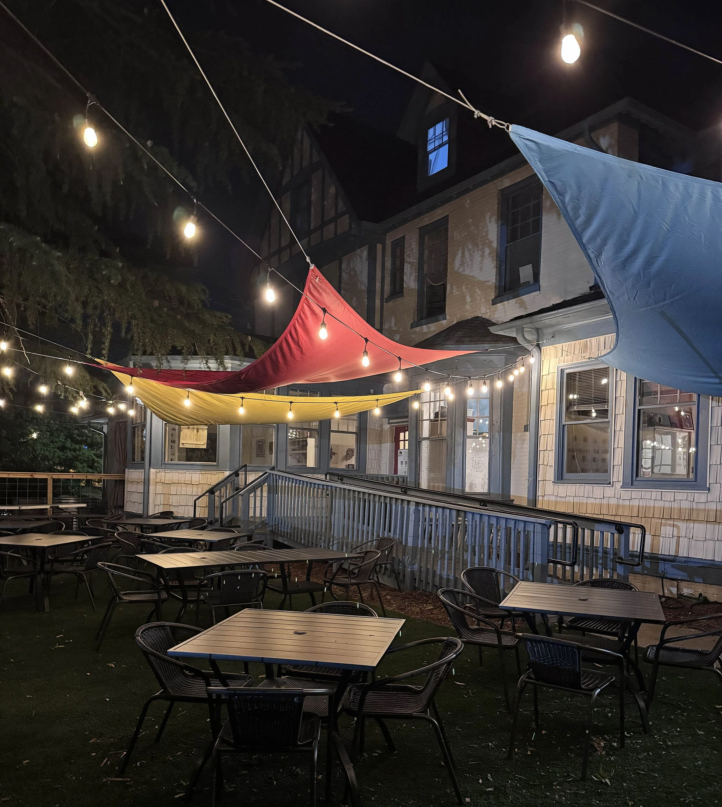 Three triangular shade sails in blue, yellow & red, illuminated by strings of Ellison lights. They're hung outside a large Tudor-style cream-colored house-turned-gallery, trimmed in blue. It's nighttime & there are tables and chairs.