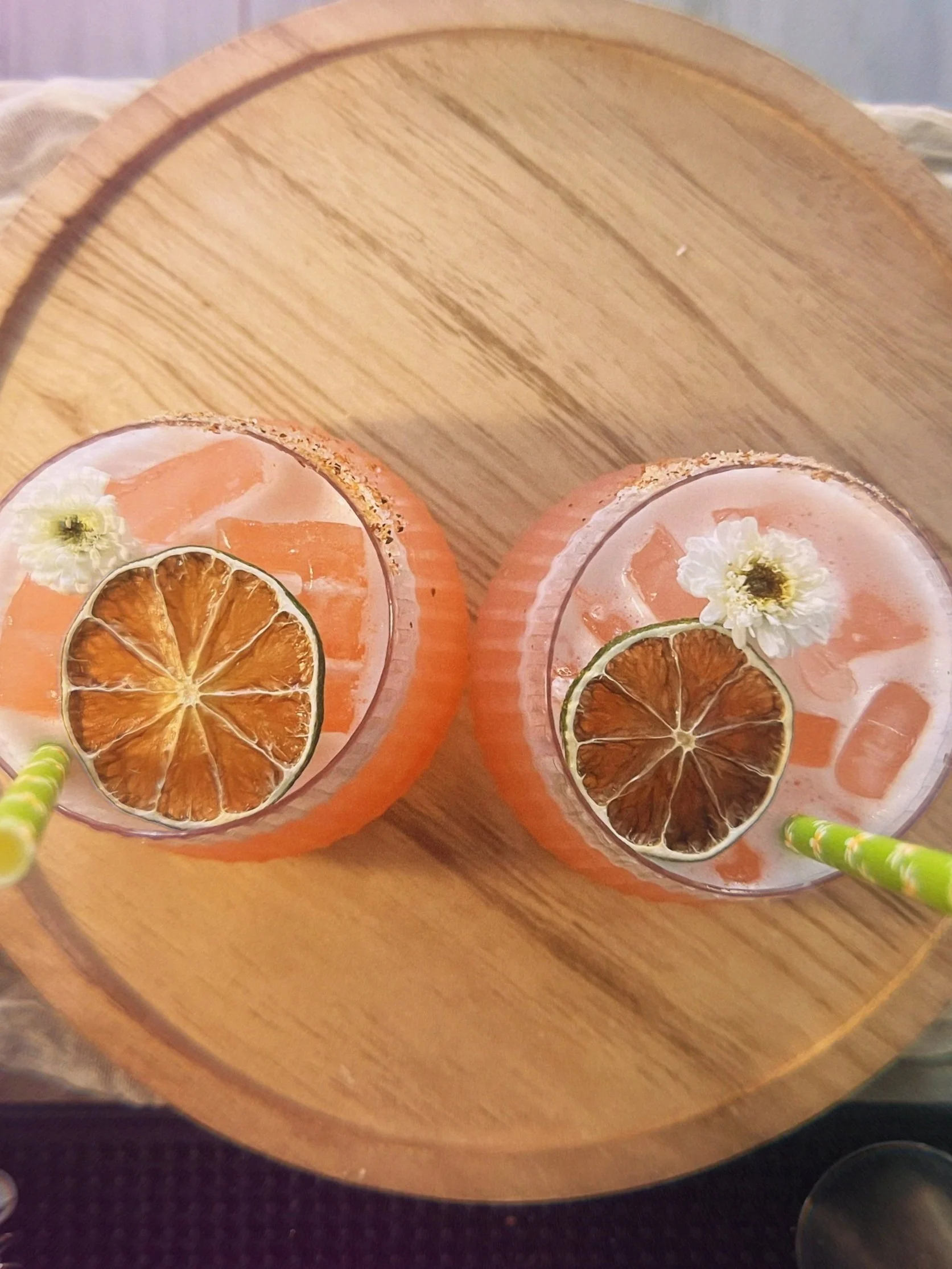 Two glasses of pink-colored cocktail garnished with dried lime wheels, small white flowers, and green striped straws, placed on a round wooden tray.