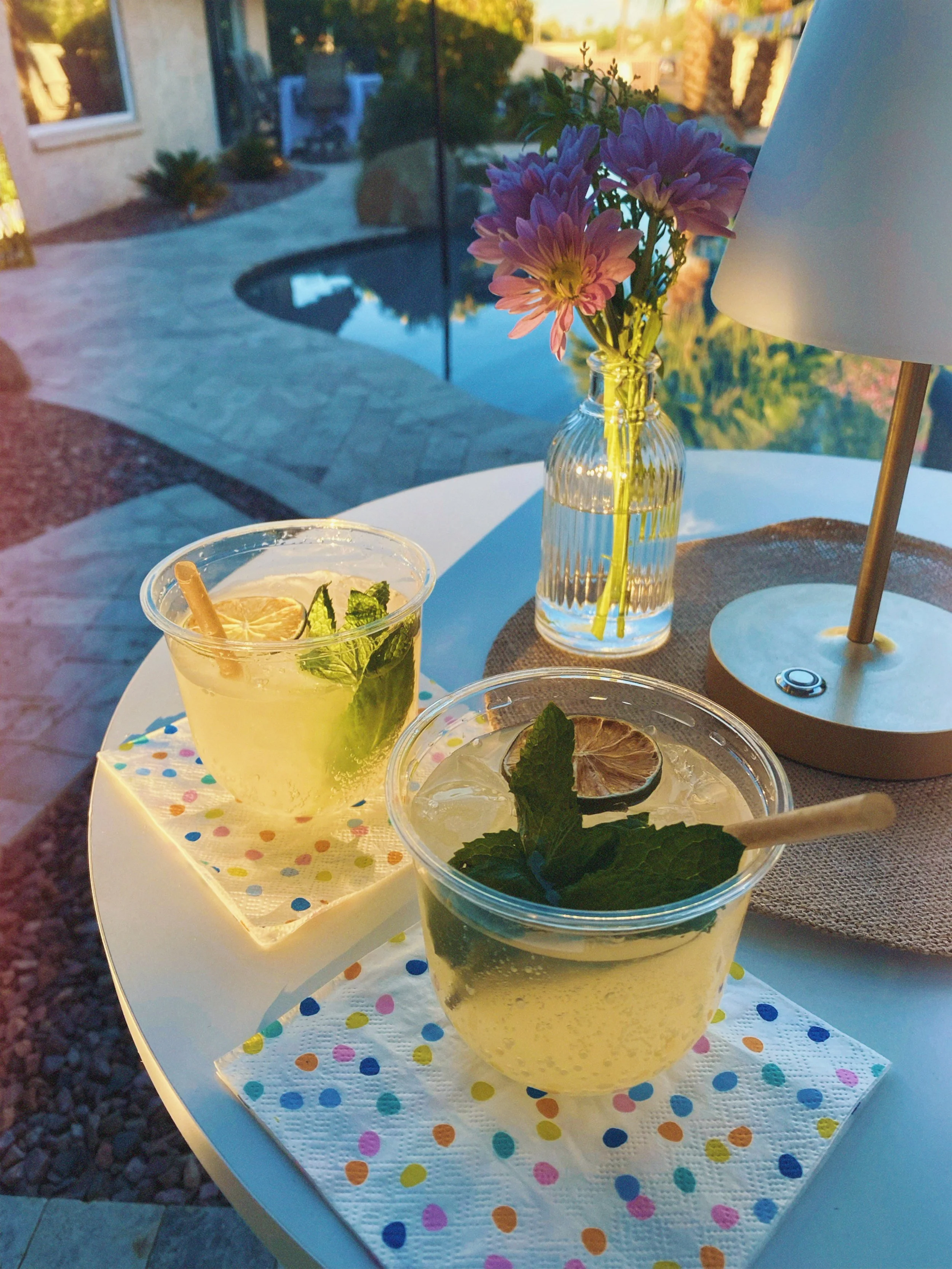 Two cocktails with lime wheels and mint leaves on a white table, a vase with pink flowers, a lamp, and a backyard pool in the background.