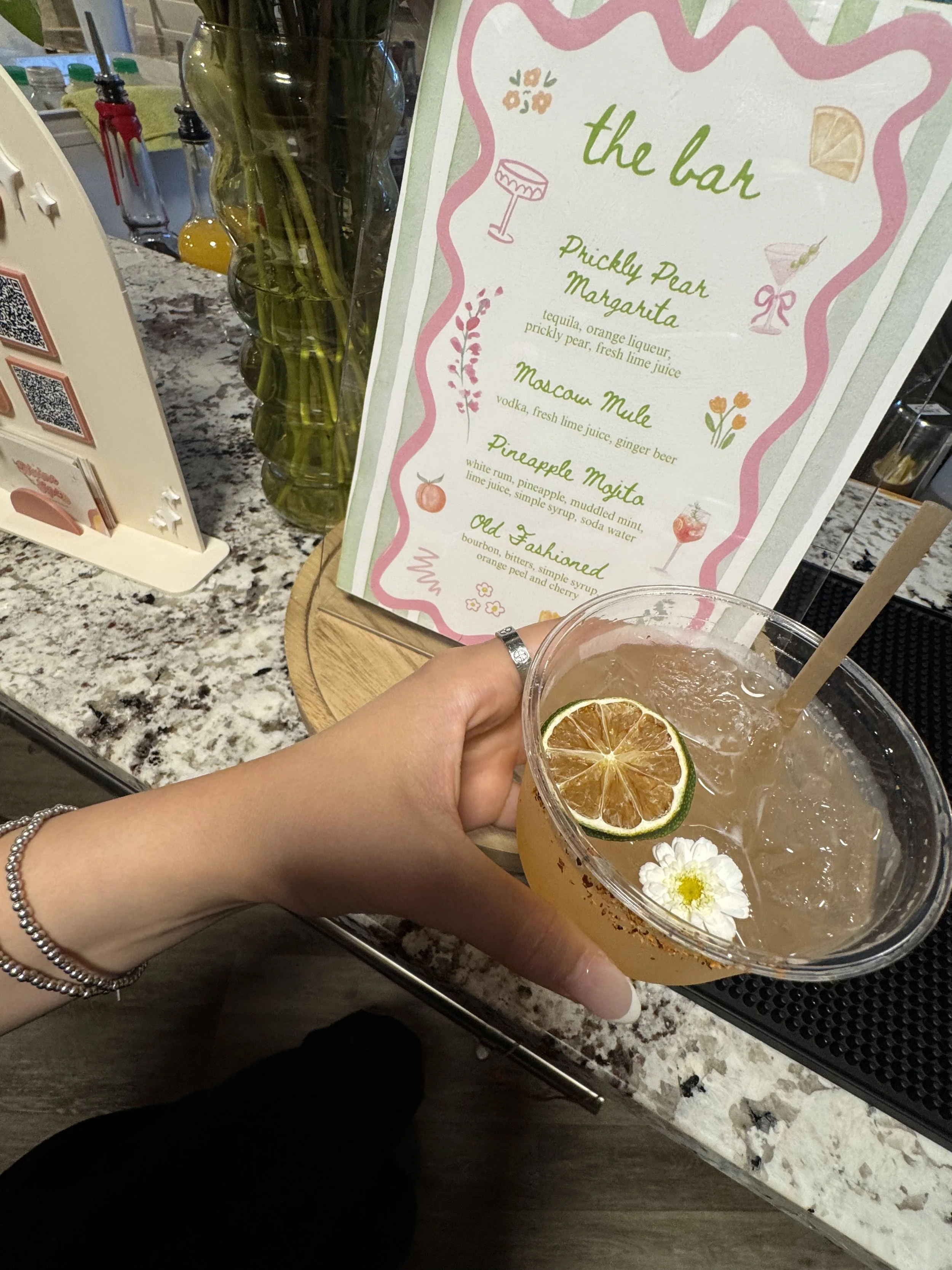 A hand holding a cocktail garnished with a slice of lime and a white flower in a clear plastic cup. In the background, a menu lists cocktails at a bar.