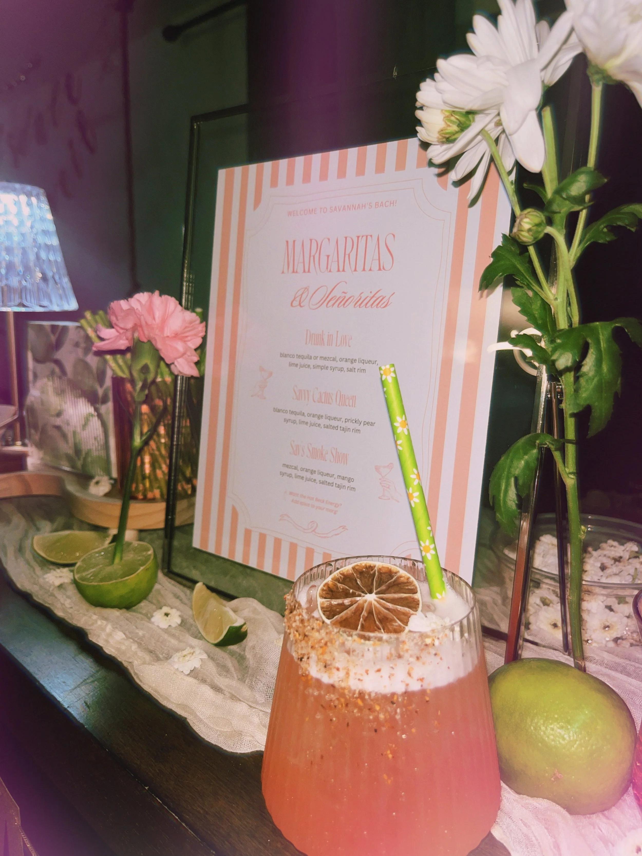A colorful cocktail with dried citrus garnish on a bar counter decorated with flowers, lime wedges, and a menu for margaritas and other drinks.