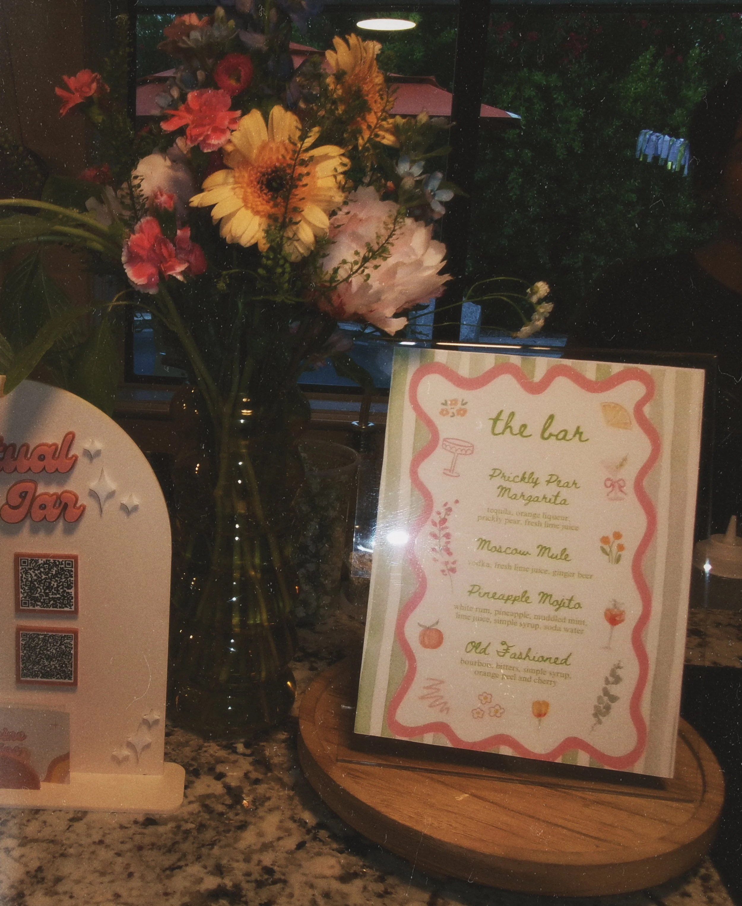 A floral arrangement of pink, yellow, and purple flowers in a dark vase next to a menu titled 'the bar' with options for drinks, on a wooden round placemat on a speckled countertop.