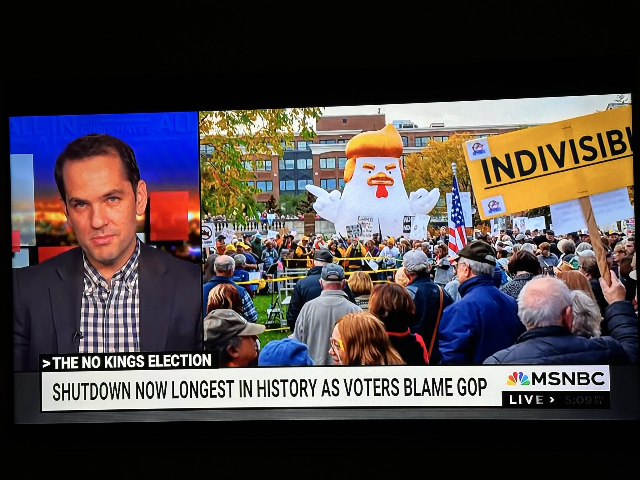 Television news broadcast showing a split-screen with a news anchor on the left and a large crowd at a protest on the right. The protest includes a large inflatables and signs, one reading 'INDIVISIBLE', with a building and fall trees in the background.
