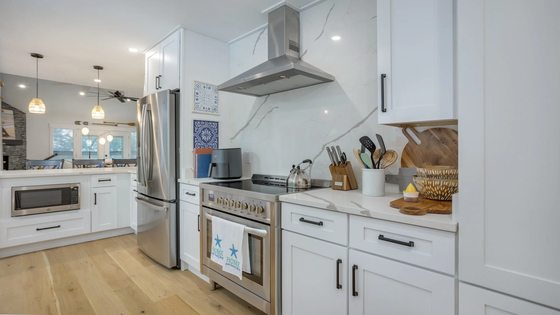 Modern kitchen with stainless steel appliances, white cabinets, and wood flooring. Features a refrigerator, stove, microwave, and decorative items like cutting boards and a utensil holder. Pendant lights and dining area visible in the background.