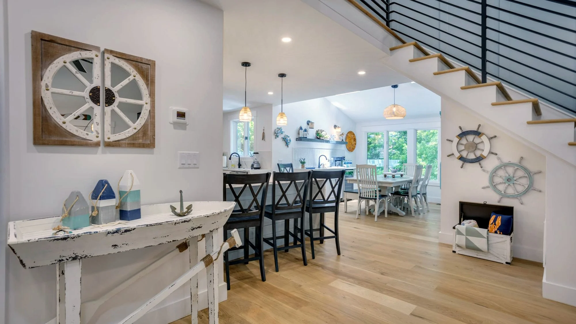 Modern coastal-themed interior with dining area, kitchen, and staircase. Nautical decor includes ship wheels and anchor. Light wood flooring and black bar stools complement the bright, open space.