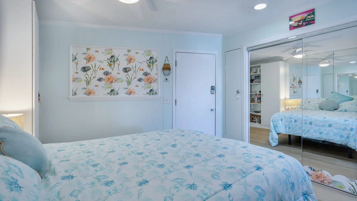 Coastal-themed bedroom with aqua bedding, mirrored closet doors, floral and fish wall art, and wooden decor. Reflections show full room layout.