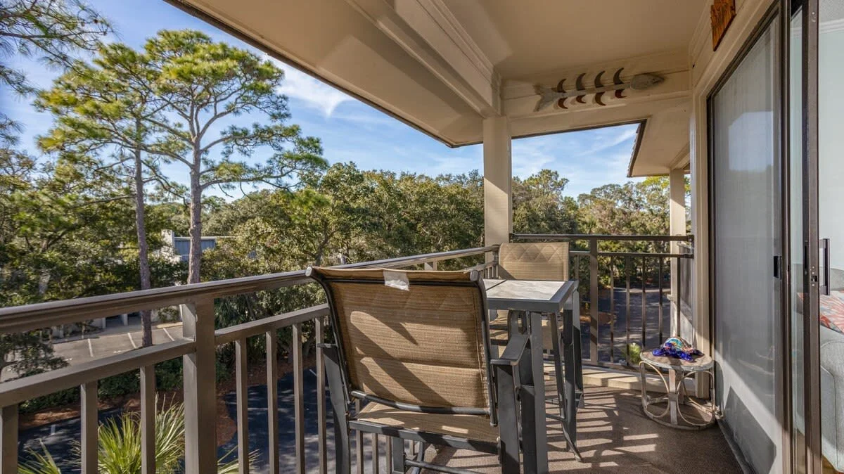 Outdoor balcony with table and chairs, overlooking trees, under a covered roof.