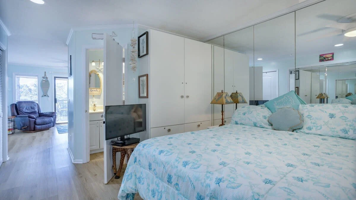 Bedroom with mirrored wardrobes, a bed with a blue and white floral comforter, bedside lamps, a small TV, and a glimpse of an adjacent bathroom and sitting area with a chair.