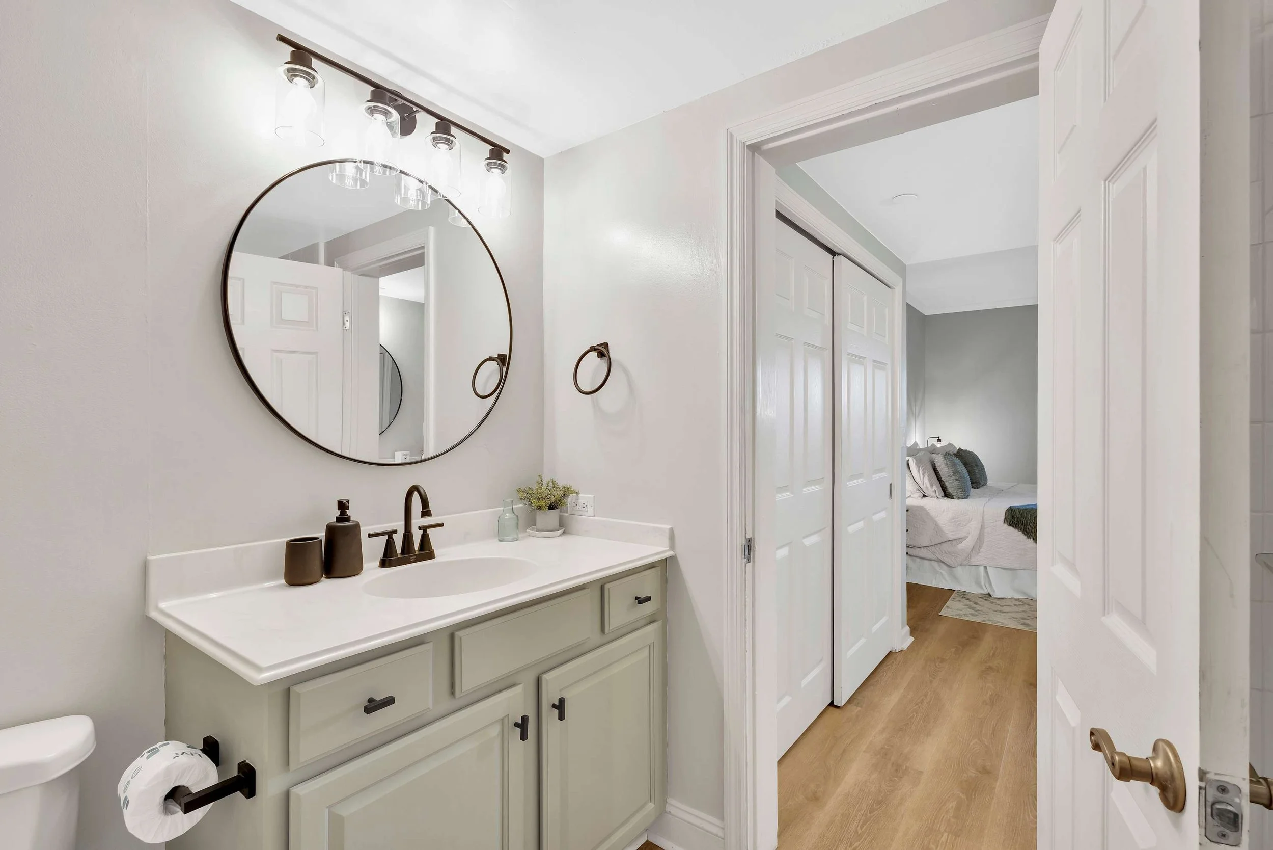 Modern bathroom with a round mirror, white vanity, black fixtures, and a view into a bedroom with a neatly made bed.