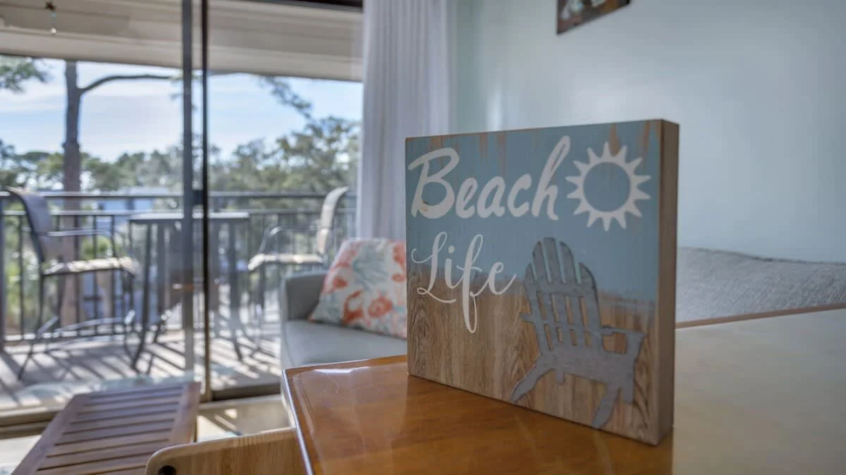 Decorative sign reading 'Beach Life' with a sun and chair design, placed on a wooden table in a room with a balcony view.