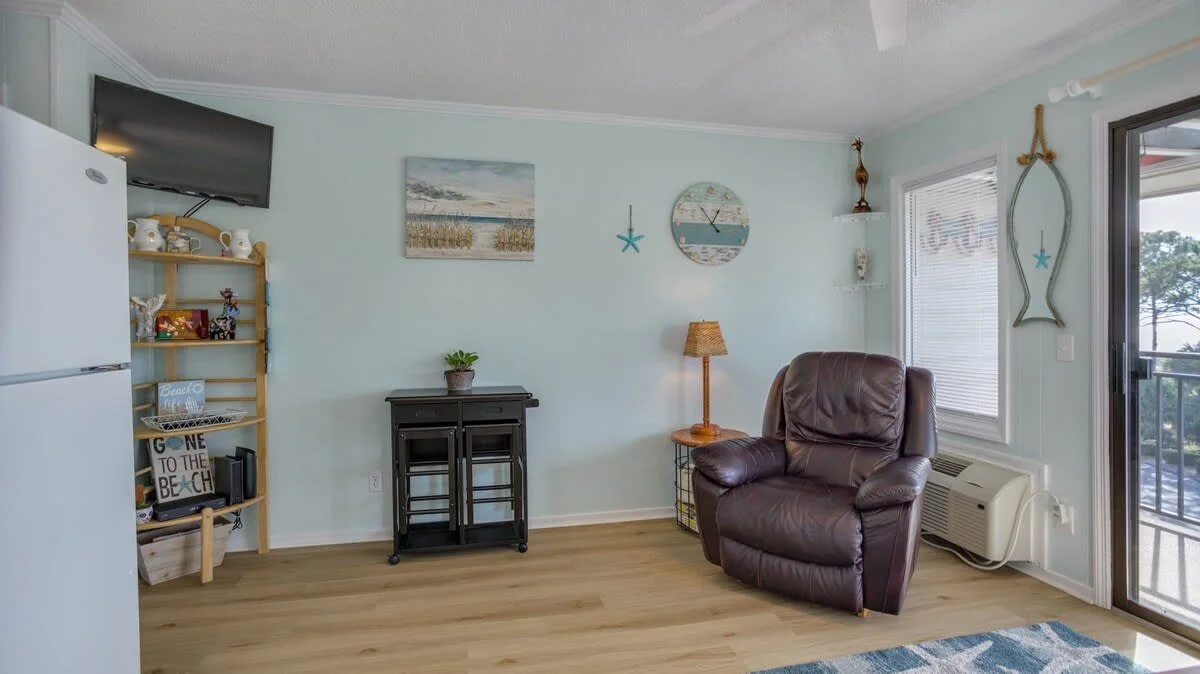 Cozy living room with a brown leather recliner, beach-themed decor, a wall-mounted TV, shelving with decorative items, a small cabinet, and a window with a view.