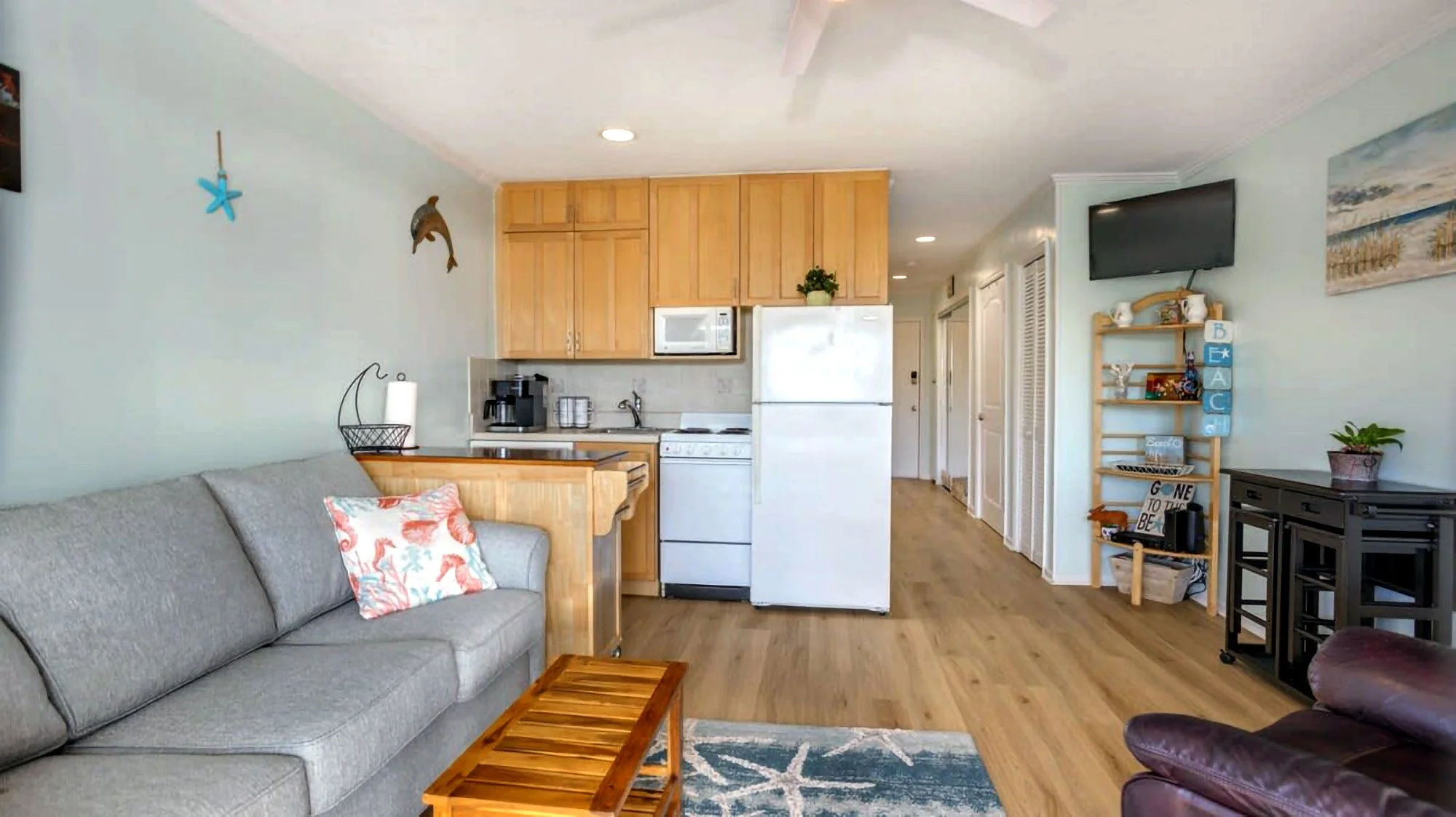 Small beach-themed apartment living room and kitchen area with light blue walls, gray sofa, wooden coffee table, and kitchen appliances like a fridge and microwave.
