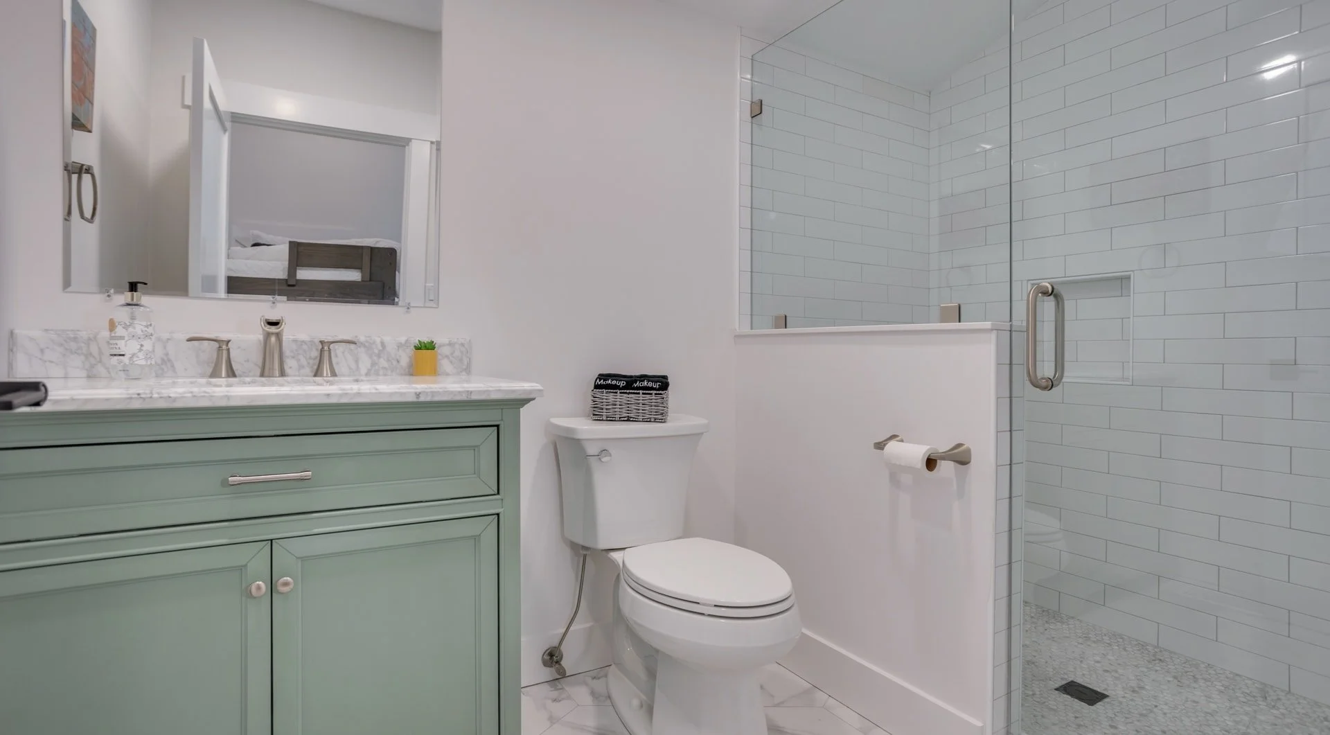 Modern bathroom with green vanity, marble countertop, toilet, glass shower with white subway tiles, round mirror, and small plant.