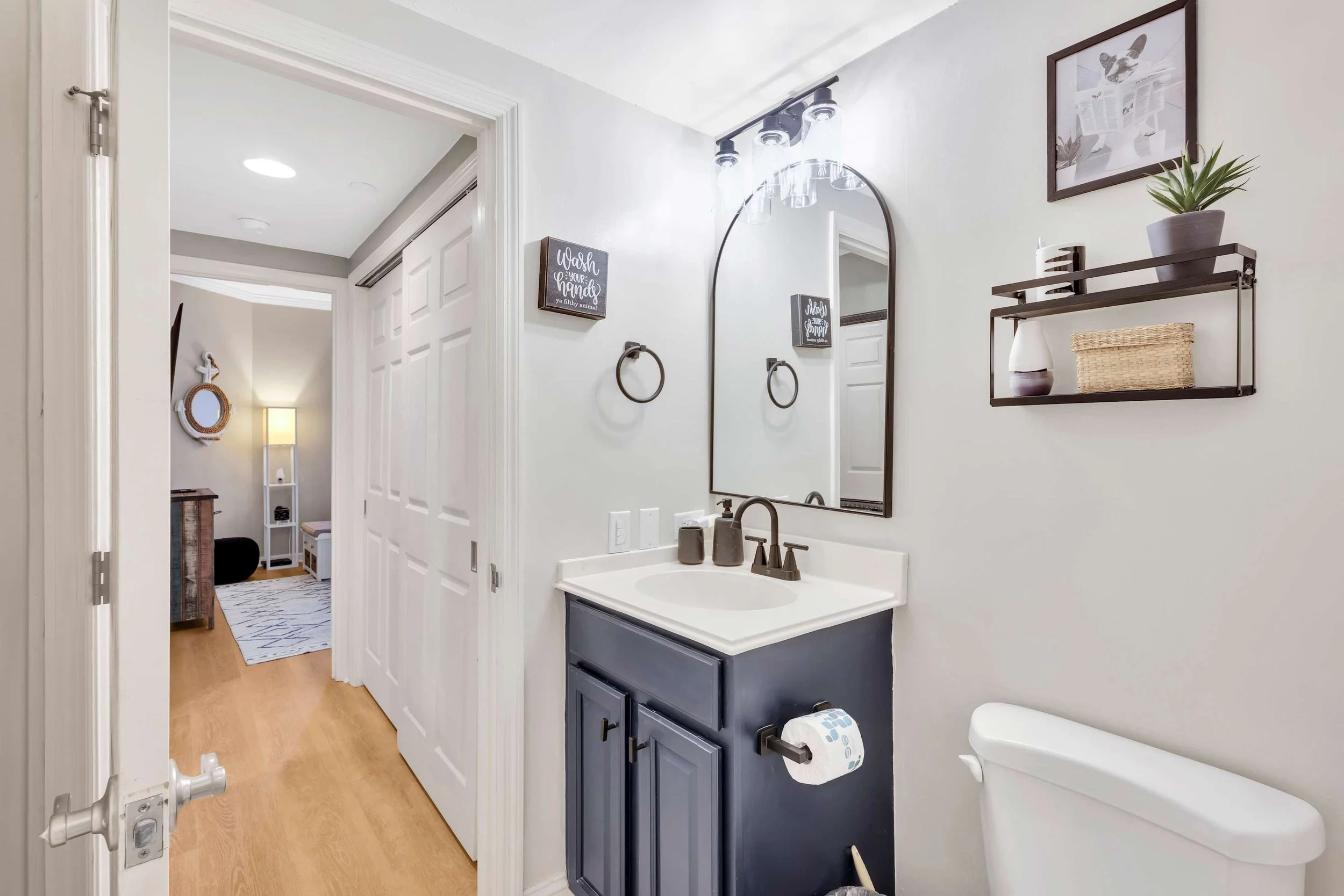 Modern bathroom with navy vanity, round mirror, and decorative shelving. Toilet paper is on a holder next to the toilet. Shelves hold a plant and a basket. Door opens to a hallway and adjacent room with wood flooring and a floor lamp.