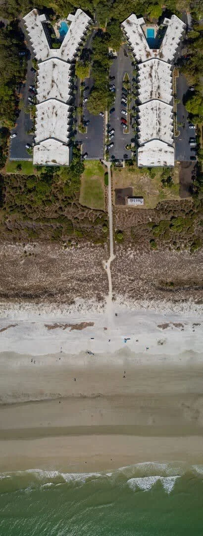 Aerial view of two large, Y-shaped apartment buildings with parking lots and a path leading through vegetation to a sandy beach along the ocean.