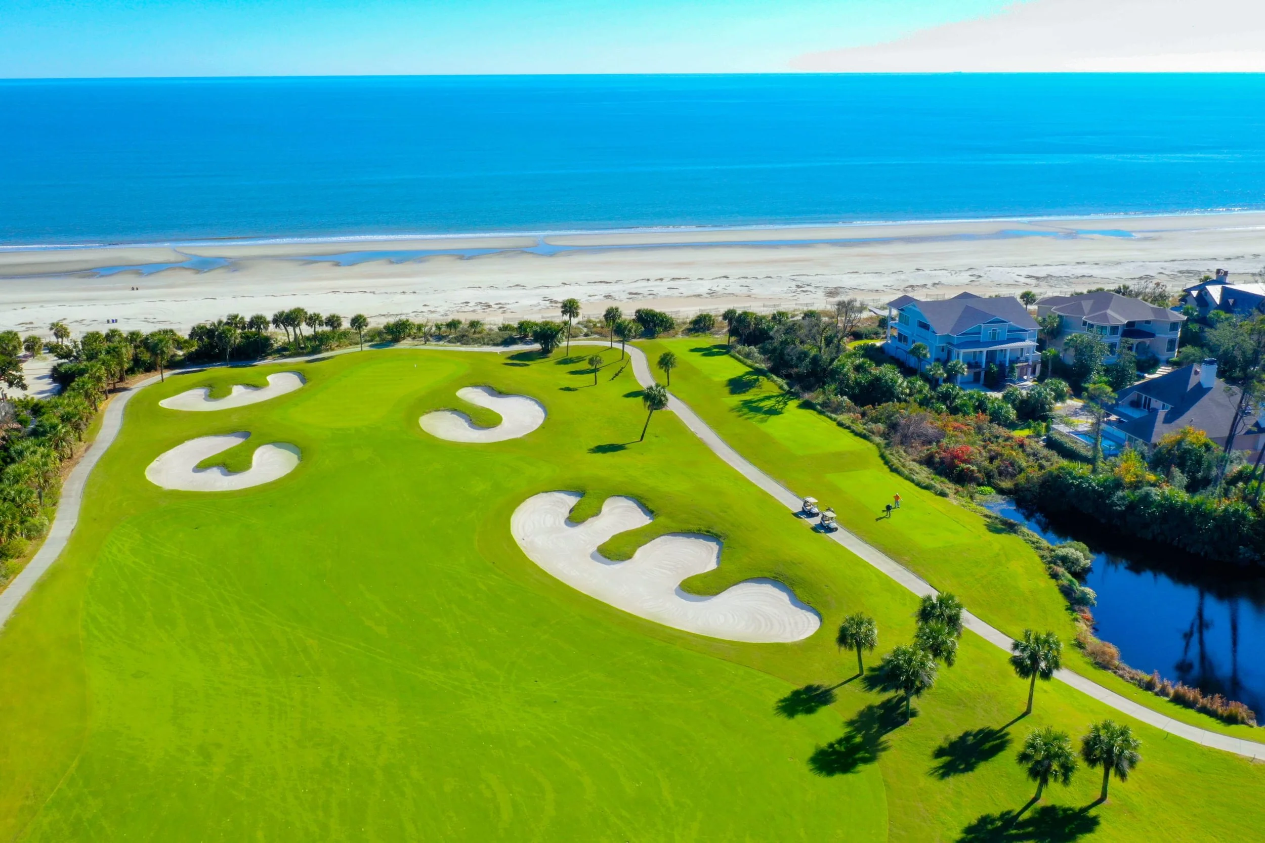 Aerial view of a seaside golf course with sand bunkers, a lush green fairway, palm trees, beach, ocean, and nearby houses.