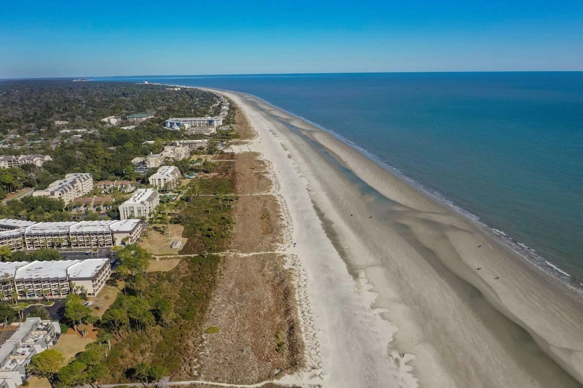 Aerial view of a long sandy beach with ocean waves on one side and buildings surrounded by trees on the other. The coastline stretches into the distance under a clear blue sky.