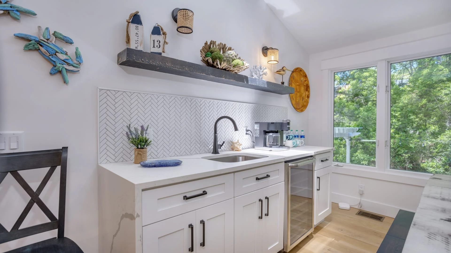 Modern kitchen nook with white cabinets and herringbone backsplash, featuring a sink, coffee maker, open shelves with decor, and a large window with trees visible outside.